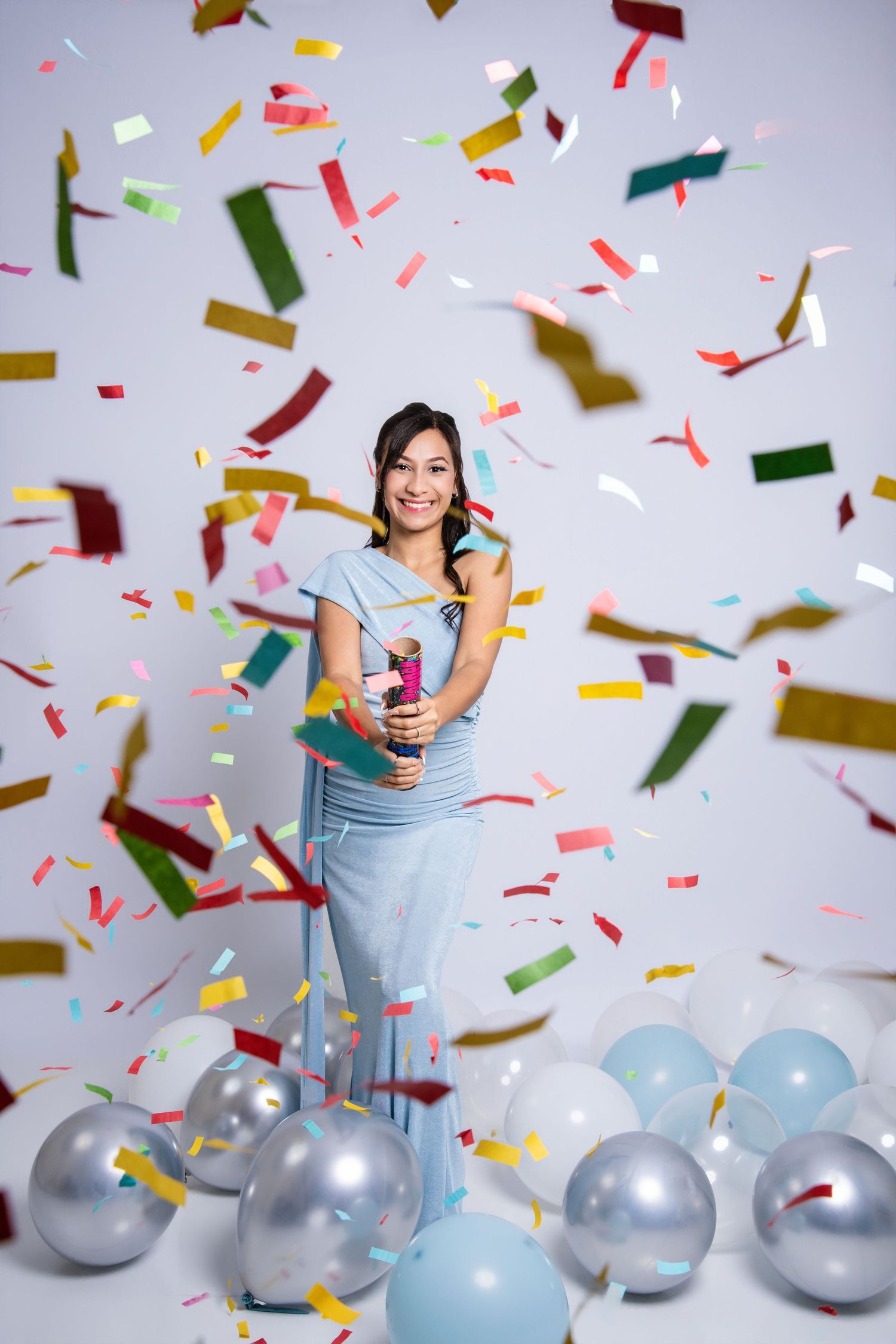 Woman in blue dress smiling while colorful confetti falls around her, surrounded by silver and blue balloons.