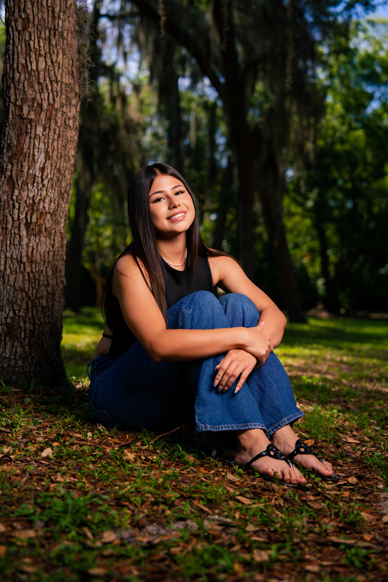 Young woman in black top and jeans sitting against tree trunk in lush green park, smiling at camera.