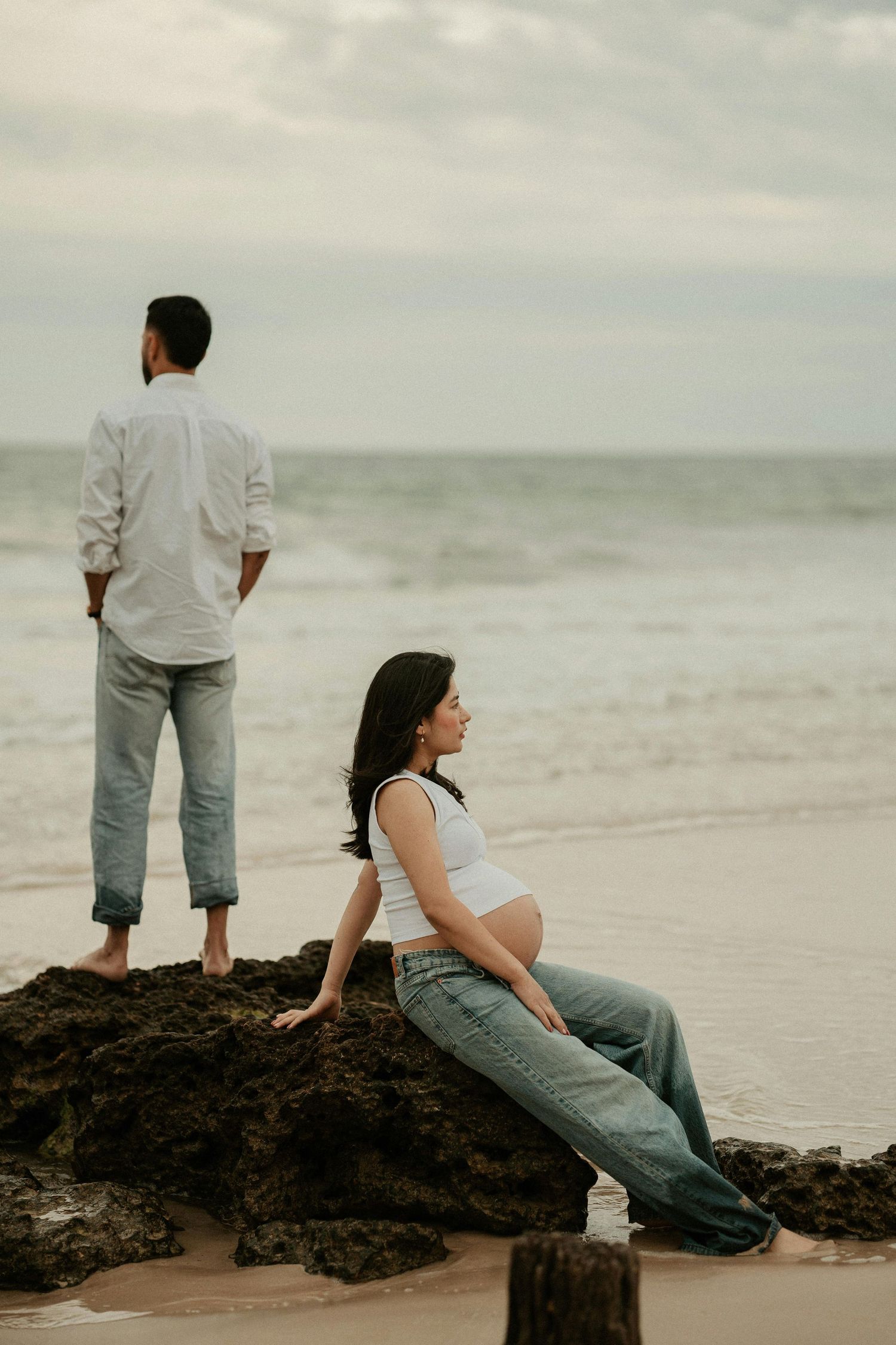 Pregnant woman in white top and jeans sits on beach rocks while man stands facing ocean during maternity photoshoot.