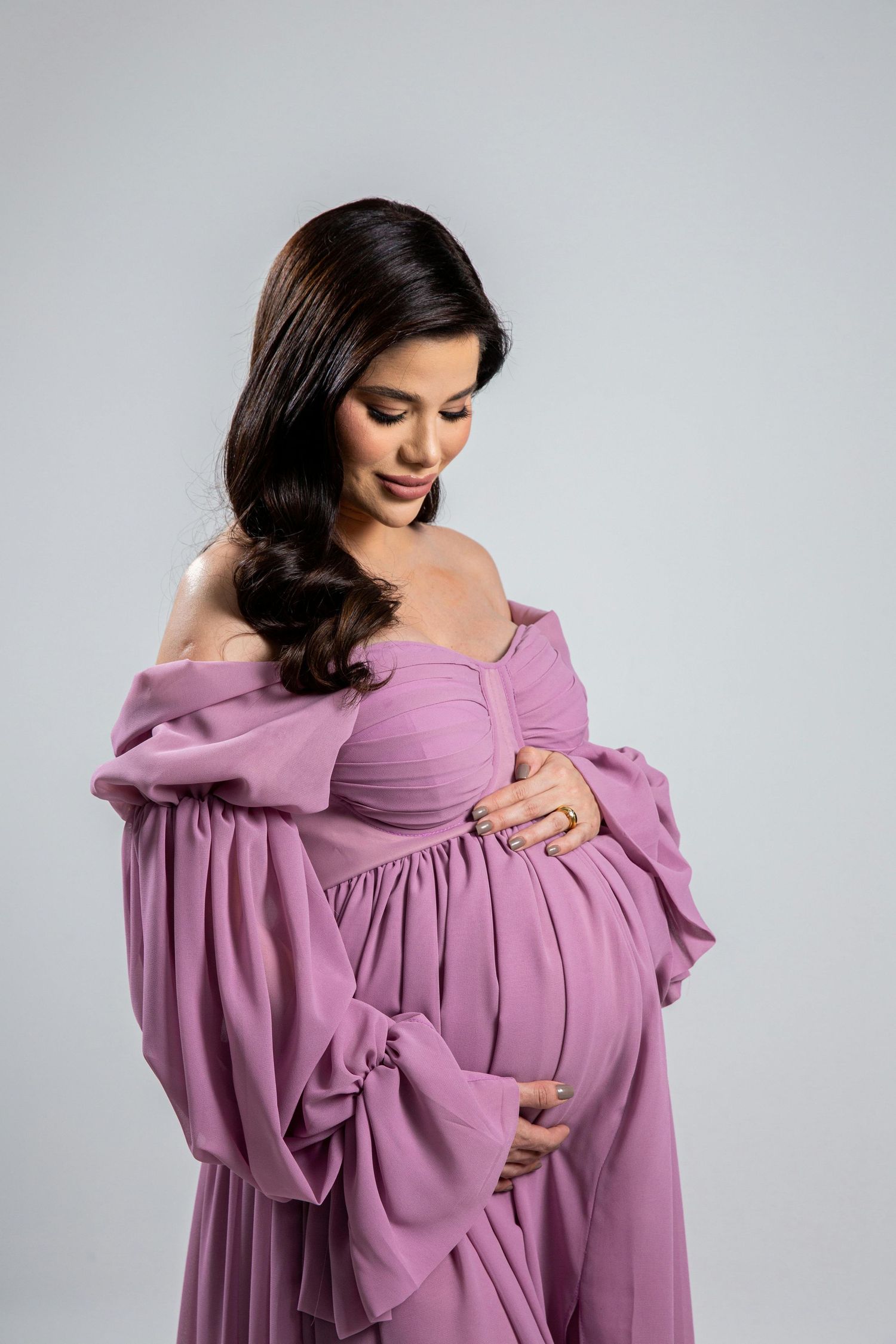 Pregnant woman in elegant off-shoulder purple maternity gown cradling her baby bump in a studio portrait.