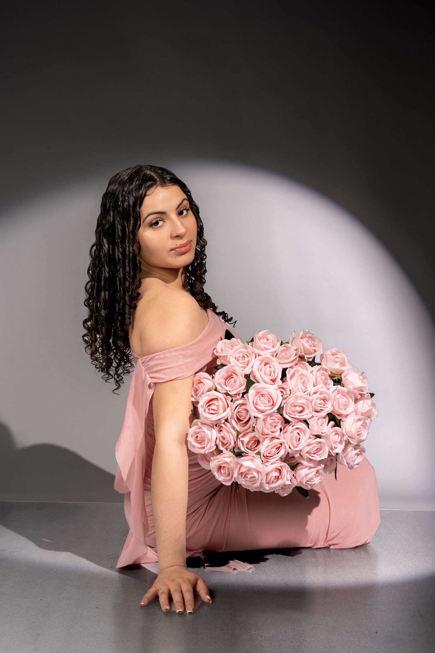 A young woman in a pink dress sits on the floor holding a large bouquet of pink roses.