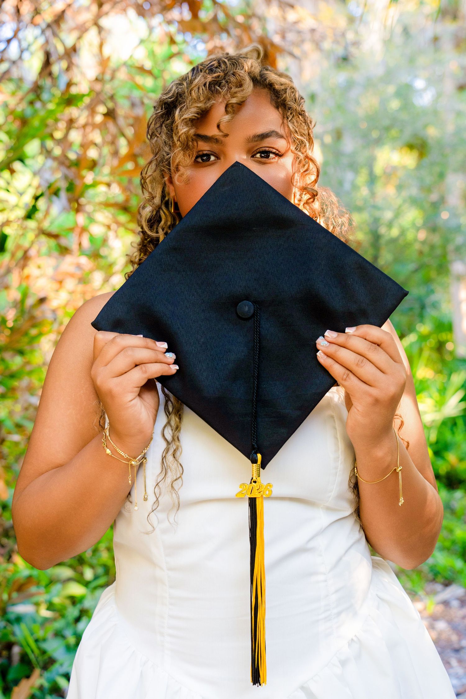 Young woman with curly hair holds black graduation cap with gold tassel in front of her face.