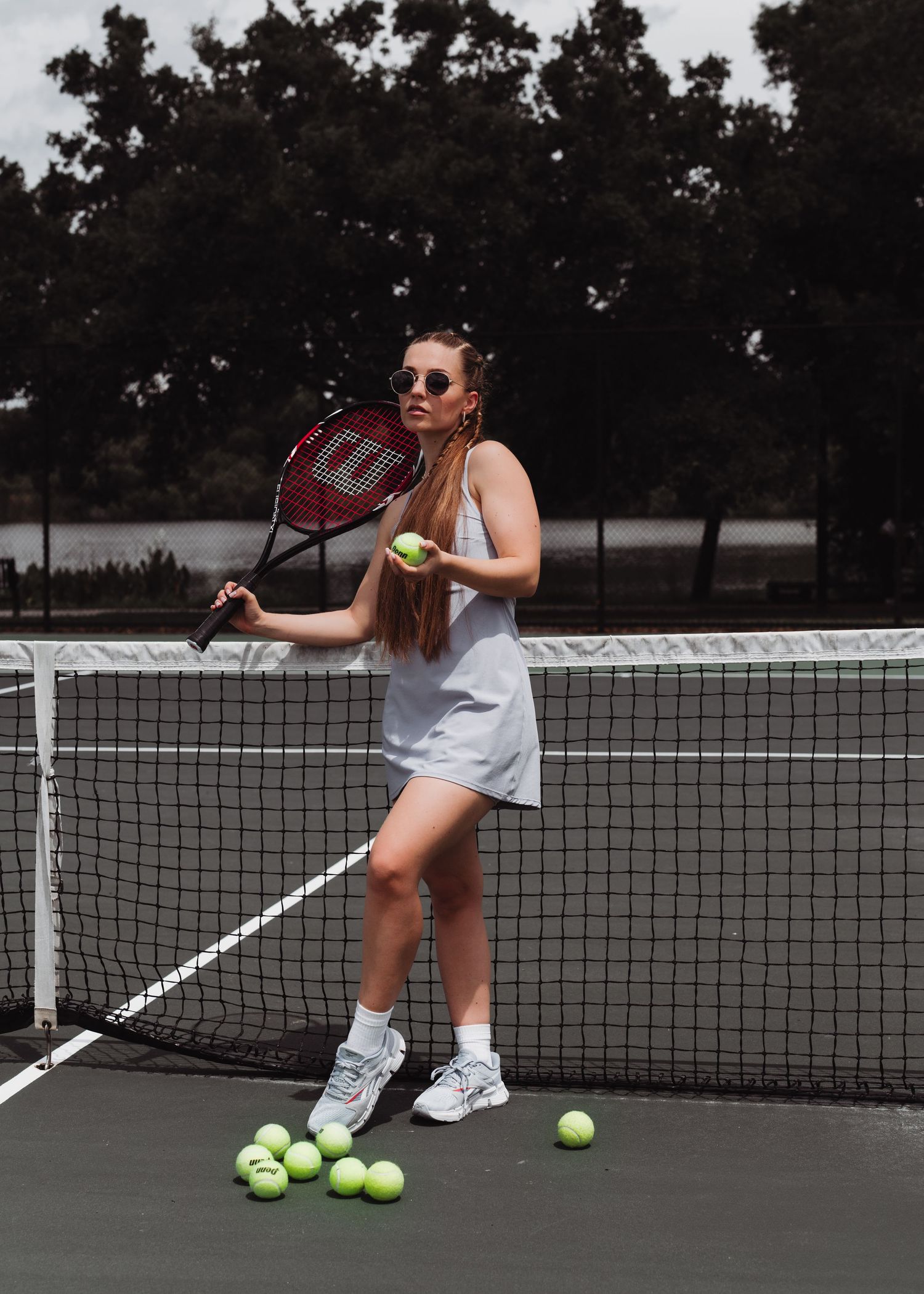 Young woman in white tennis dress holding Wilson racket at net on outdoor court.