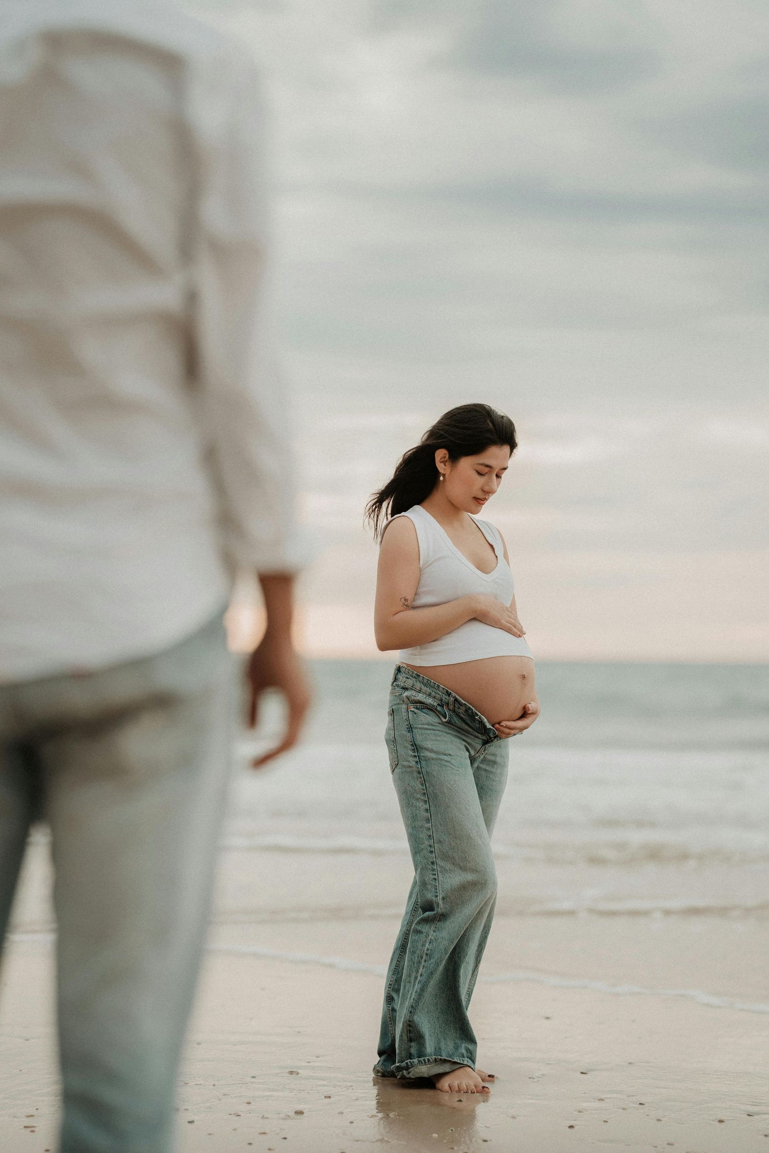 Pregnant woman in white crop top and jeans cradling her baby bump on a beach at sunset.