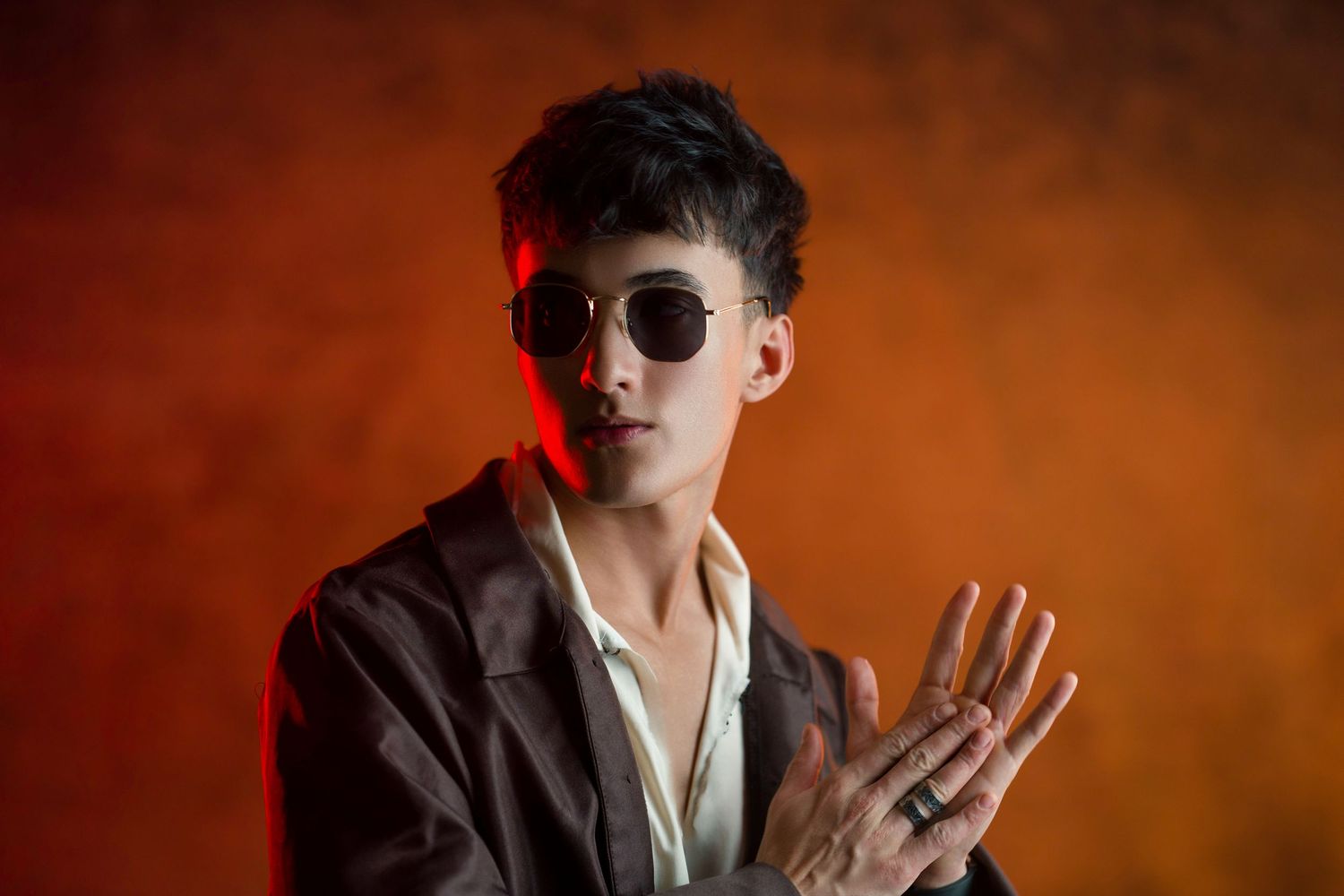 Stylish young man in sunglasses and brown blazer poses against dramatic orange background.