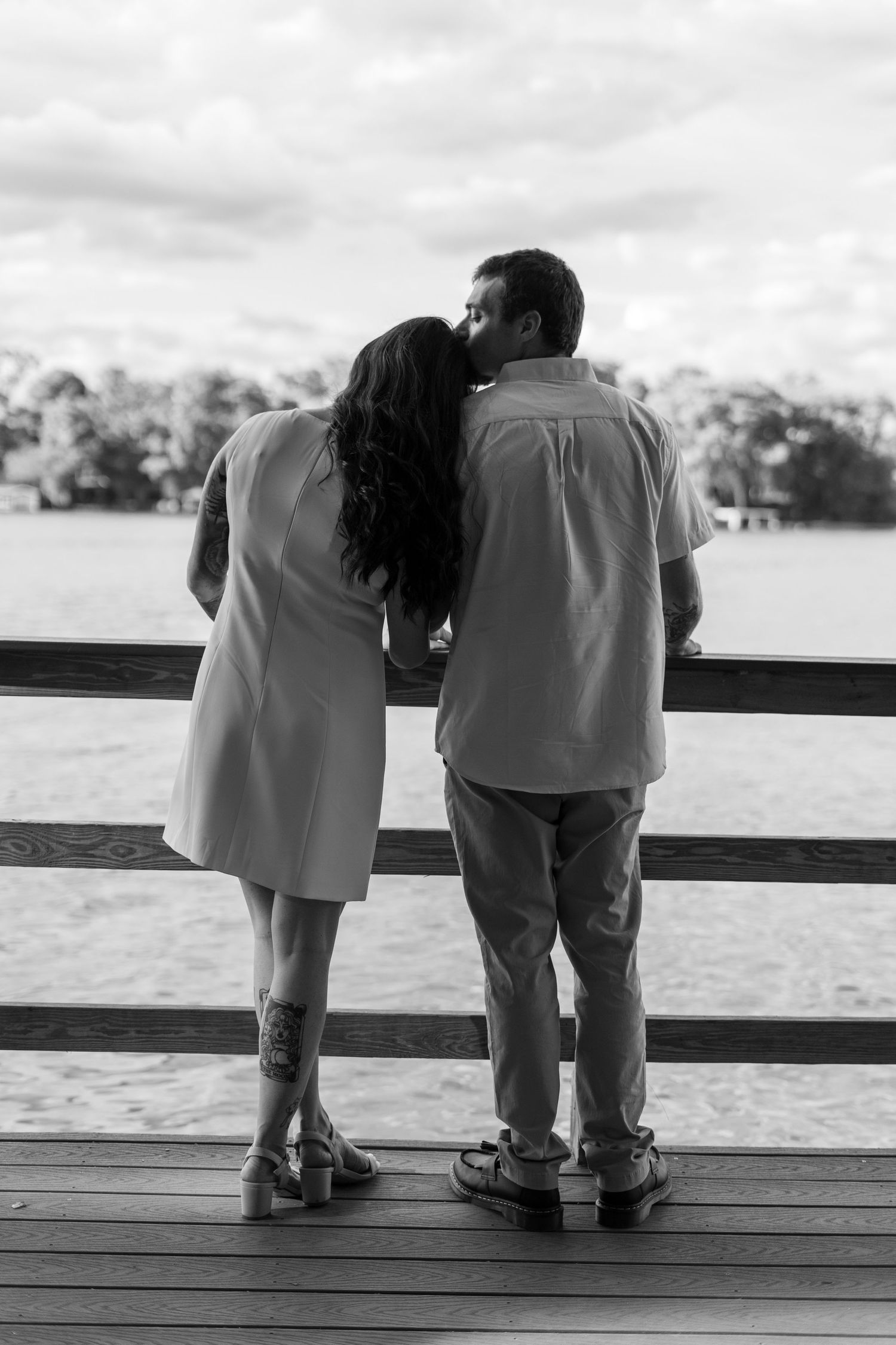Black and white photo of a couple kissing on a wooden dock overlooking a serene lake surrounded by trees.
