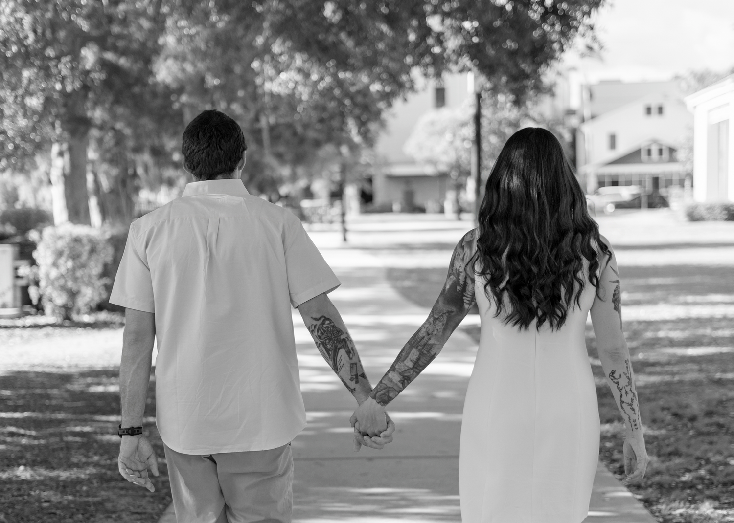 Black and white photo of a tattooed couple holding hands while walking on a sidewalk in a park.