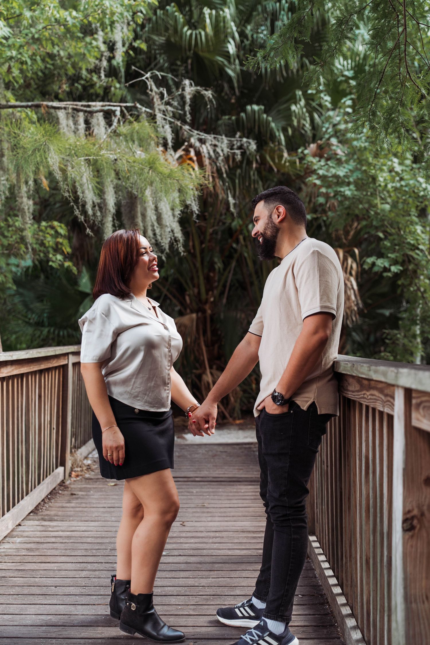 Young couple holding hands and smiling at each other on wooden bridge surrounded by green foliage.