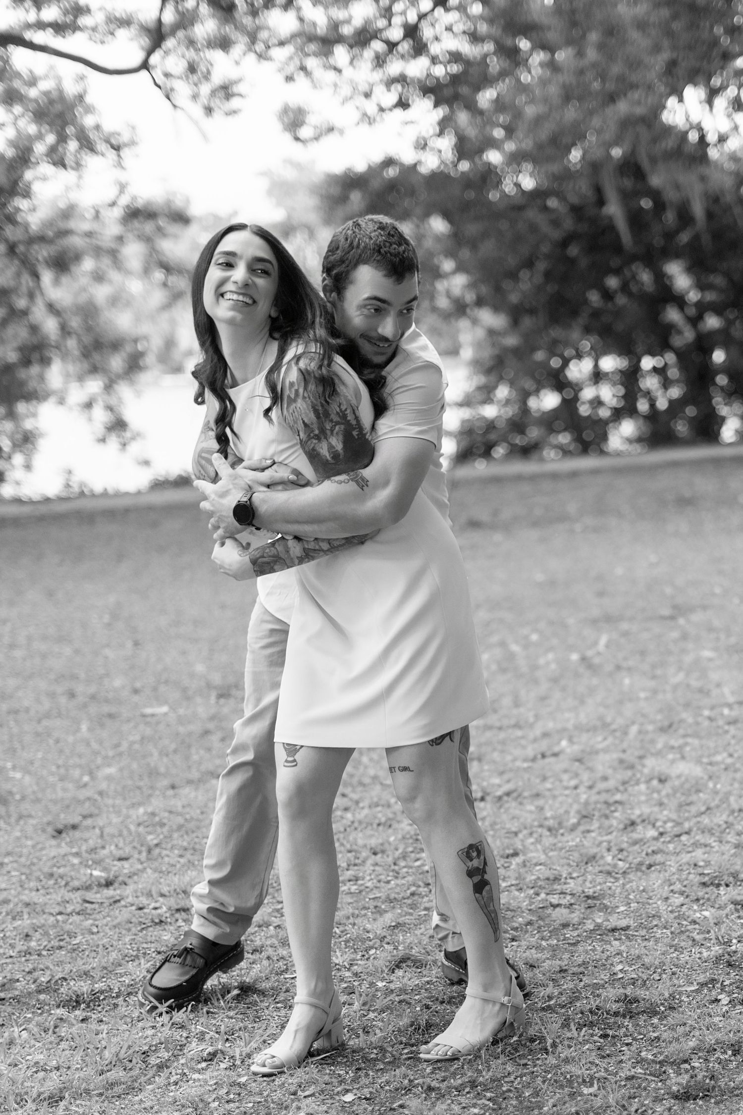 Black and white photo of a happy couple embracing in a park, man hugging woman from behind, both smiling outdoors.