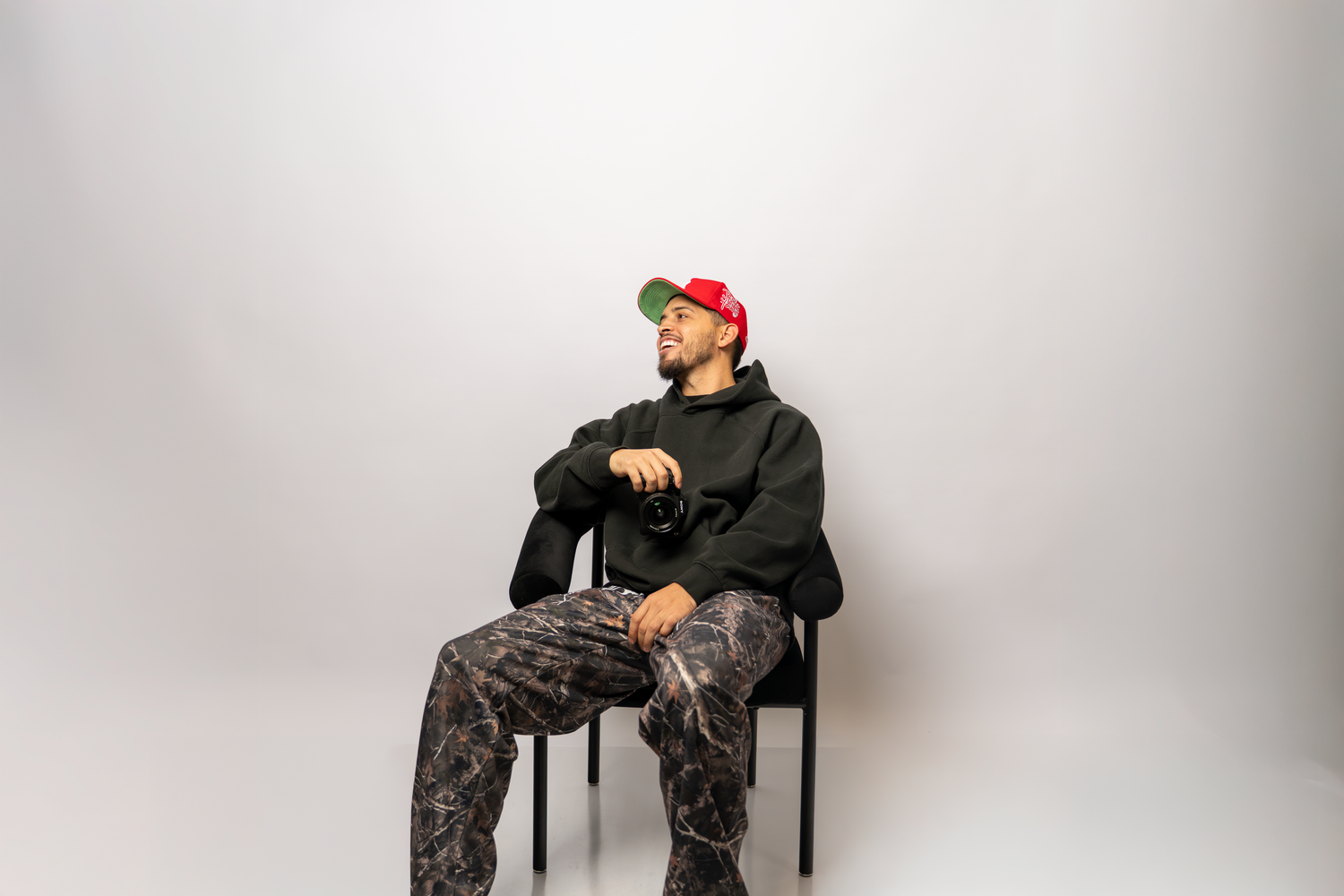 A young man in a colorful cap and patterned pants sits on a chair against a white background.
