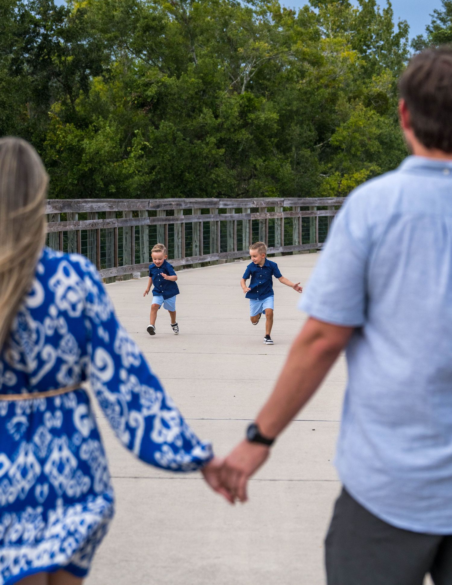 Two young boys running toward parents holding hands on a bridge with green trees in background.