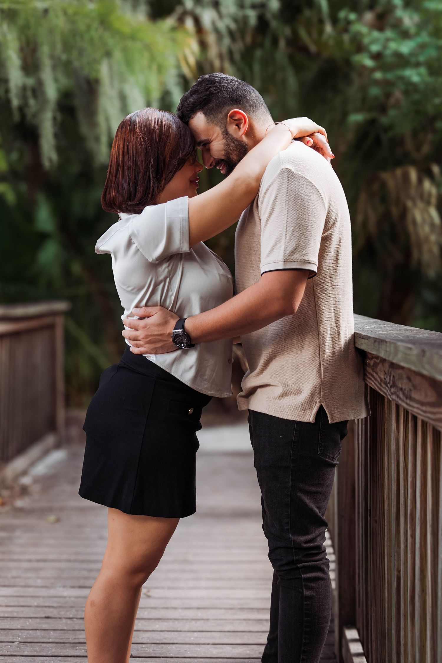 A couple shares a romantic embrace on a wooden bridge surrounded by lush greenery.