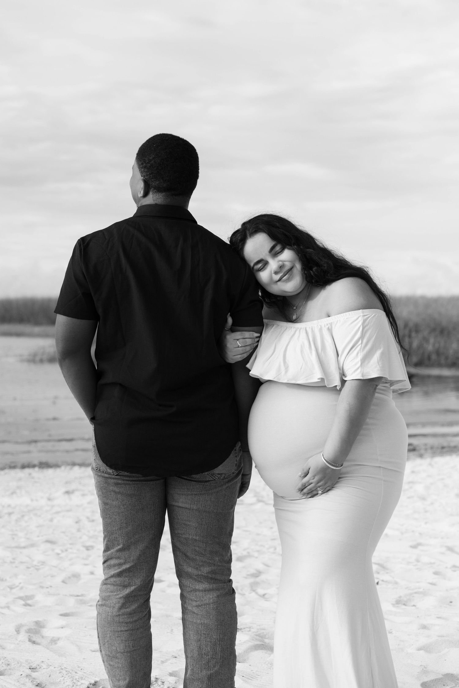 Black and white maternity photo of pregnant woman in white dress leaning on partner at beach.