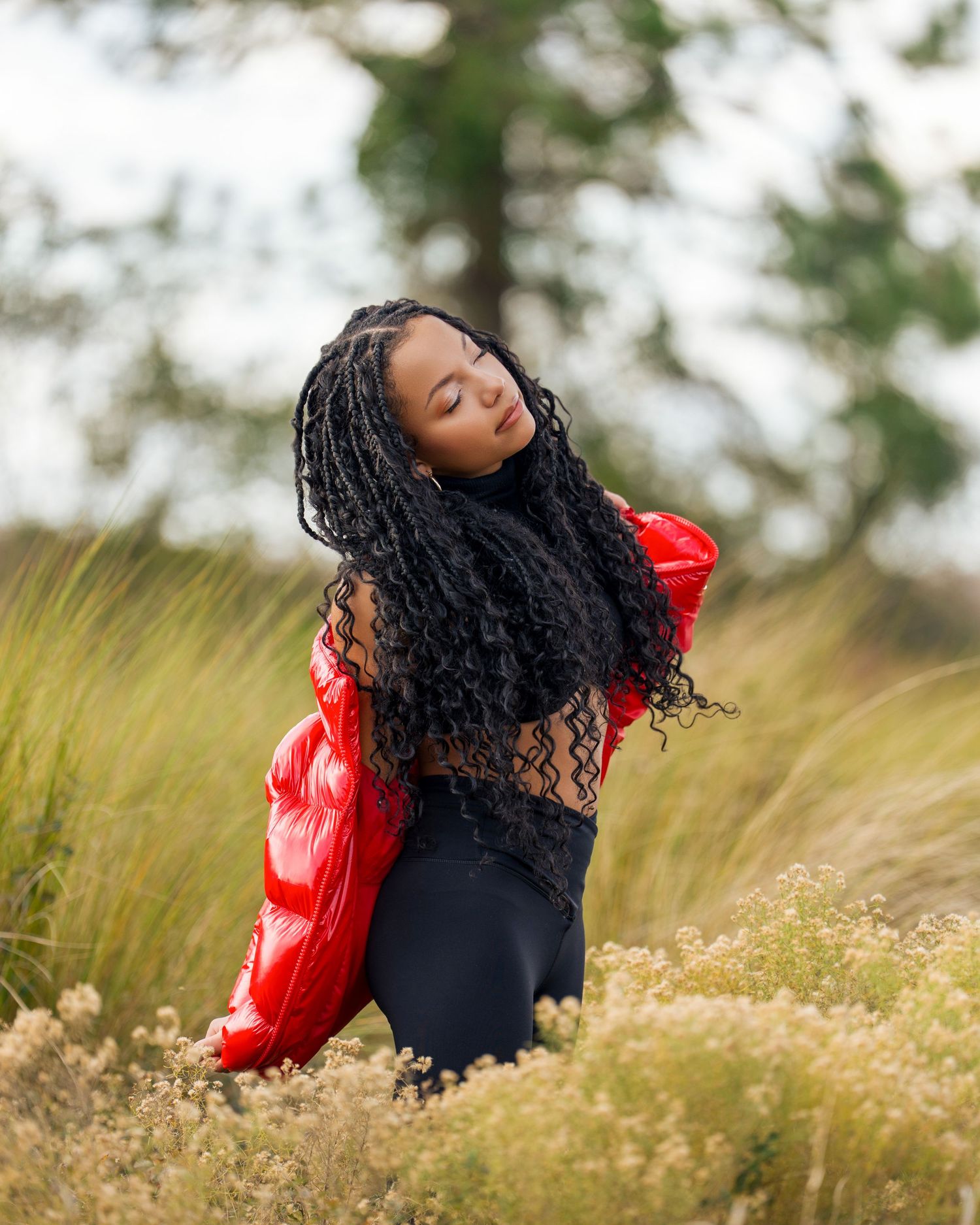 Woman with long black braids poses in a field wearing a red puffer jacket and black outfit.