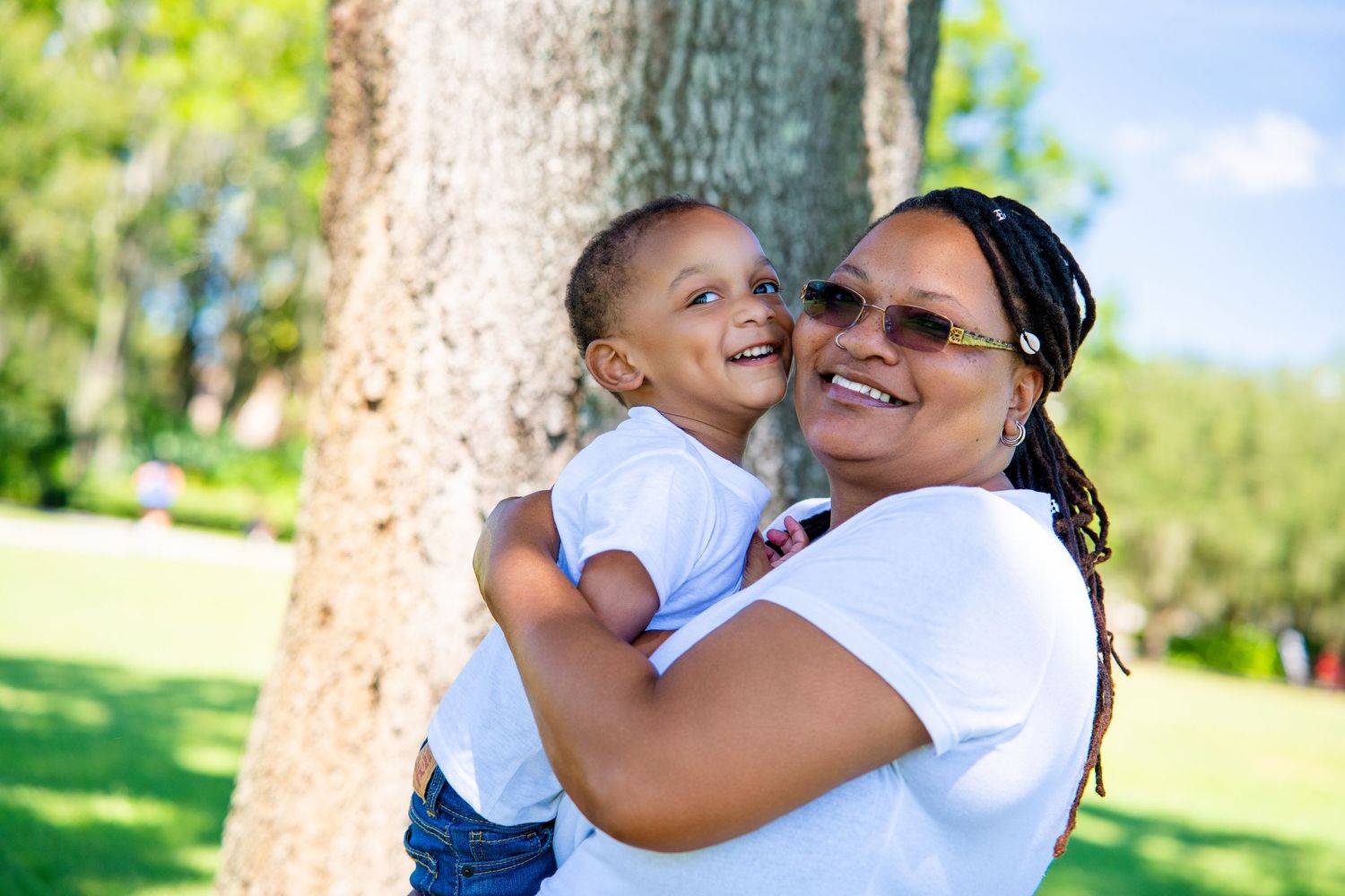A smiling mother holds her happy young child outdoors near a tree in a sunny green park.