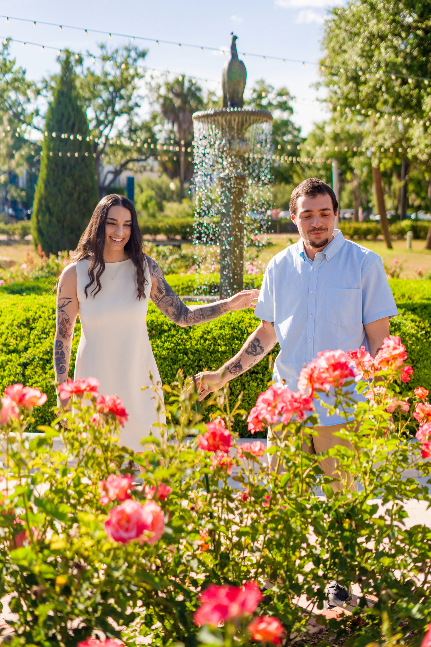 Couple with tattoos standing among pink roses near a peacock fountain in a lush green park on a sunny day.