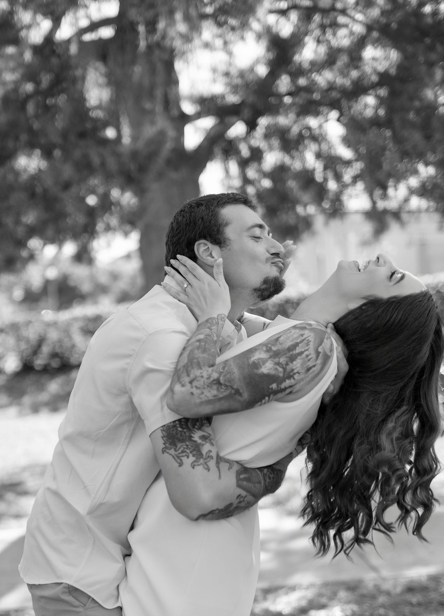 Black and white photo of a tattooed couple sharing a romantic embrace outdoors near a large tree.