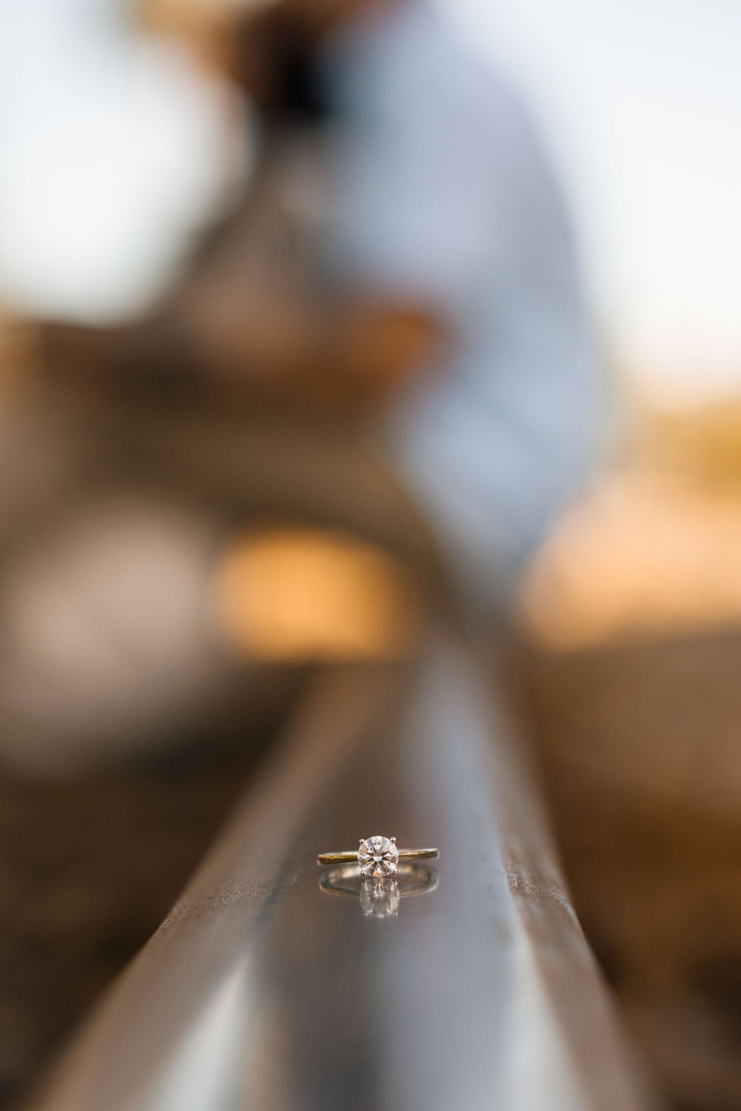 Diamond solitaire engagement ring balanced on railroad track with blurred couple in background.