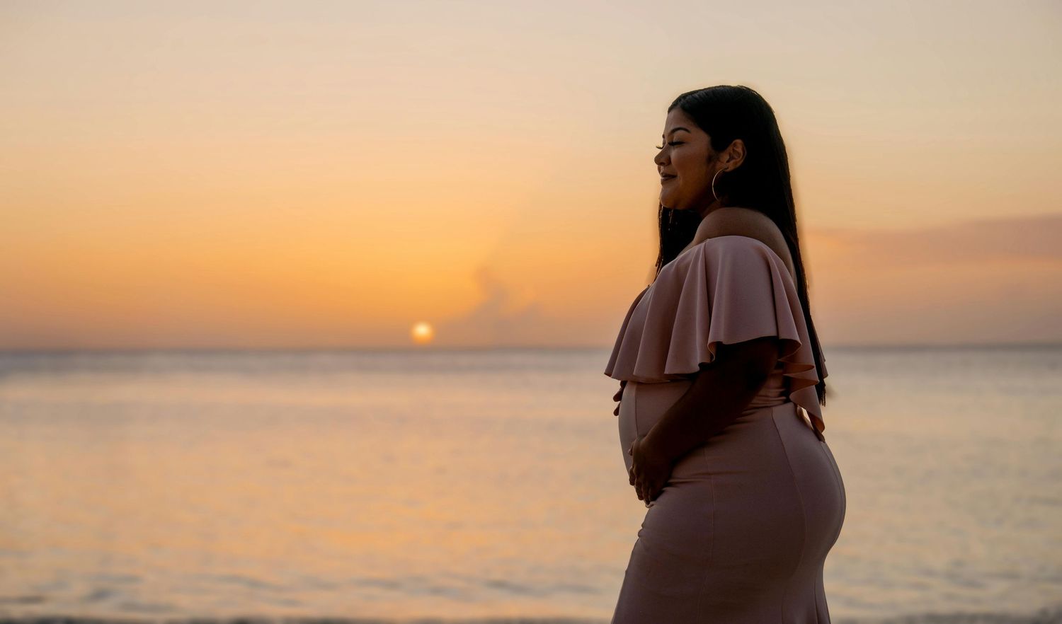 Pregnant woman in beige dress standing on beach at golden sunset, cradling baby bump by ocean.