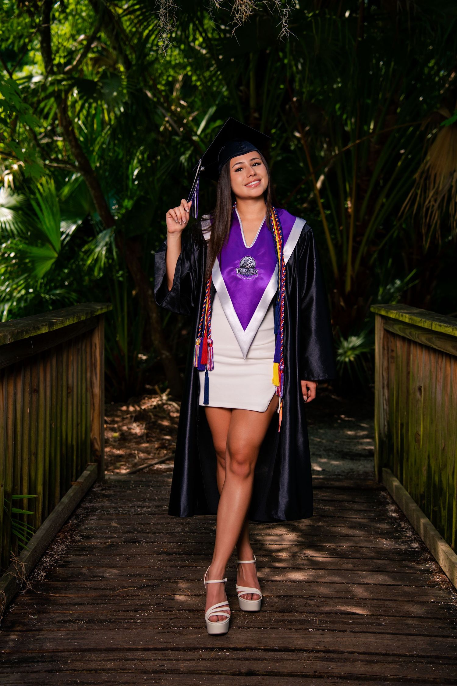 Smiling graduate in black cap and gown with purple stole and honor cords poses on wooden bridge.
