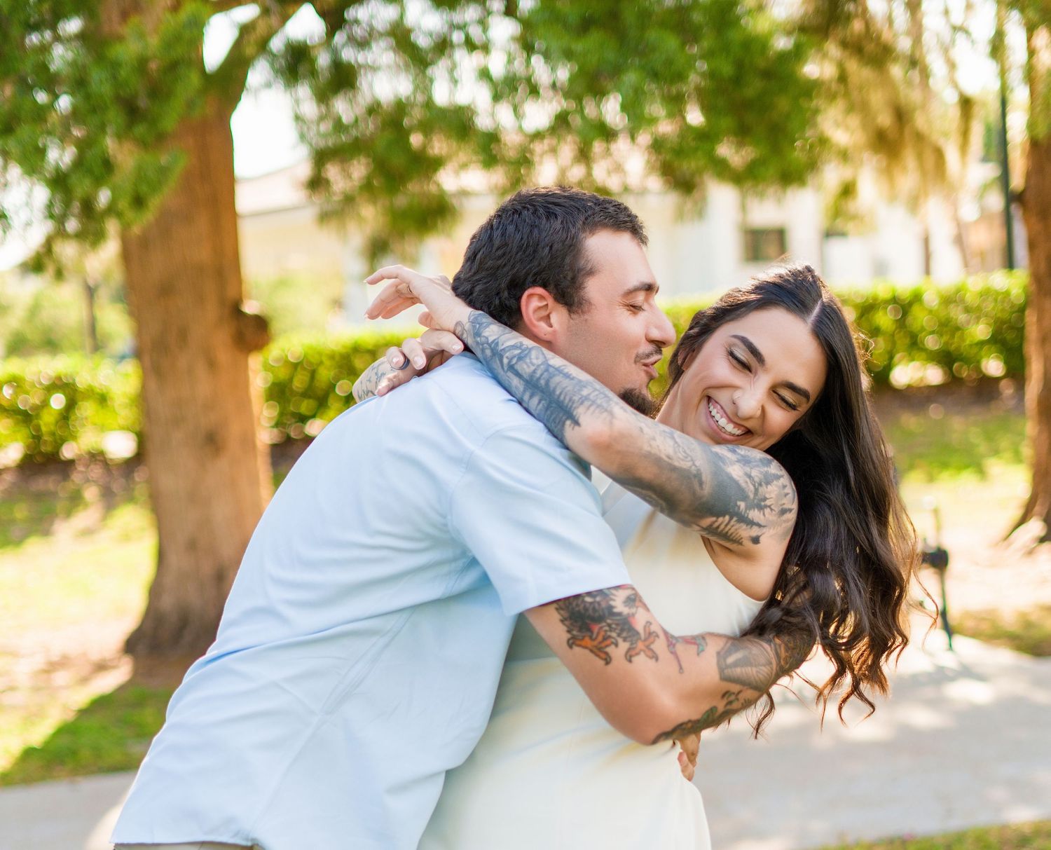 Couple embracing outdoors, tattooed man hugging smiling woman with long dark hair near green trees.