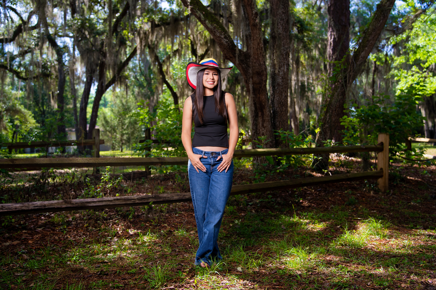 Young woman in colorful hat and jeans poses by wooden fence under moss-draped oak trees in lush green park.