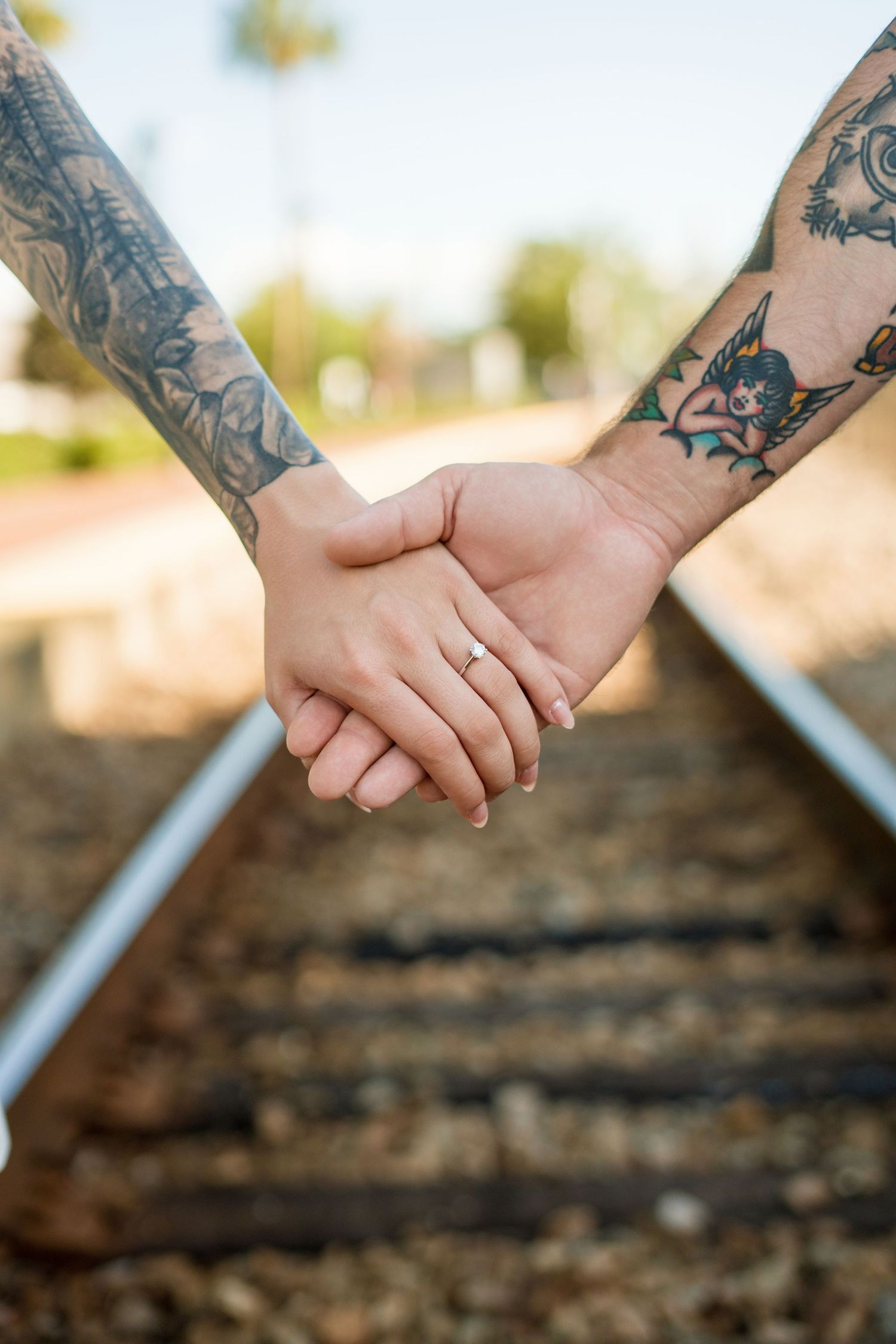 Tattooed couple holding hands on railroad tracks, woman wearing diamond engagement ring, close-up shot outdoors.