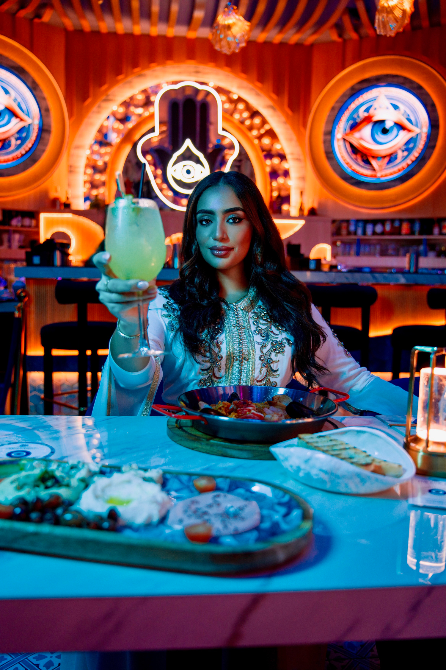 Woman raising a cocktail glass at a vibrant Middle Eastern restaurant with neon Hamsa and eye decor.