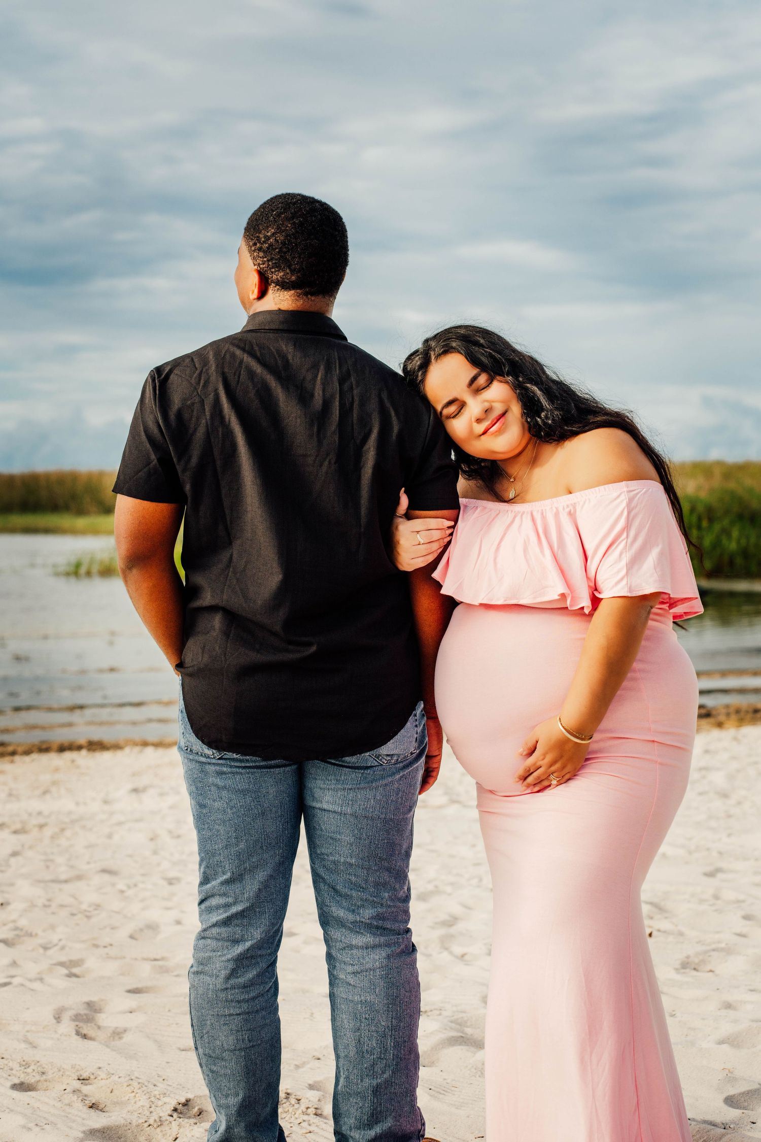 Pregnant woman in pink off-shoulder gown leans on partner's shoulder at sandy beach near water.