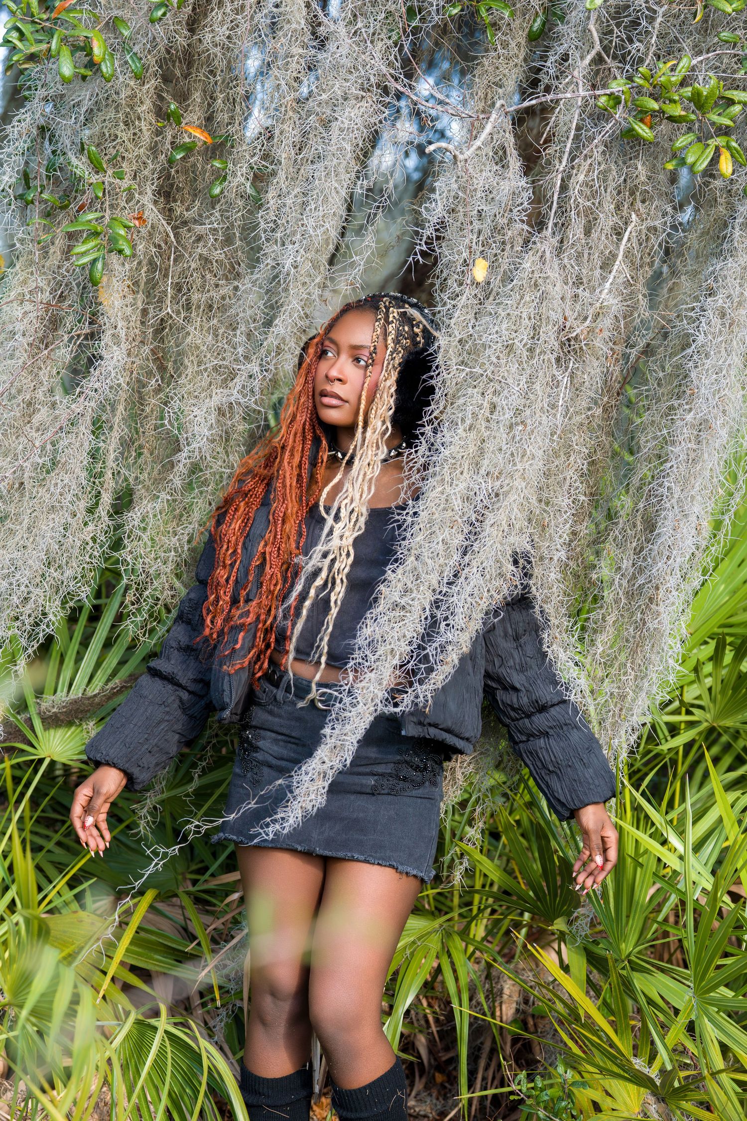 Young woman with orange locs standing among Spanish moss hanging from trees in nature.