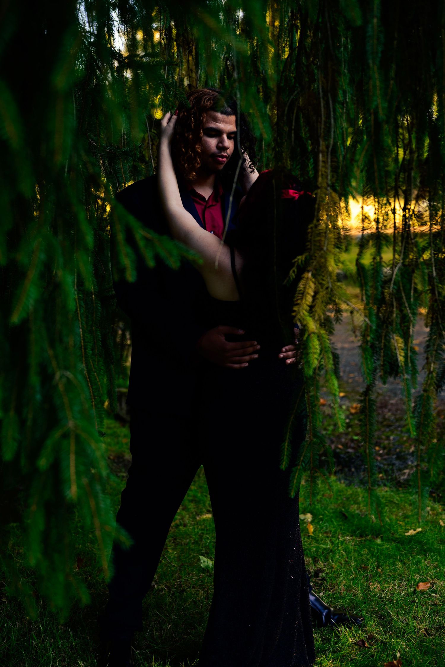 A couple embraces romantically under a weeping willow tree at dusk in a lush green park setting. High school homecoming event neo OH