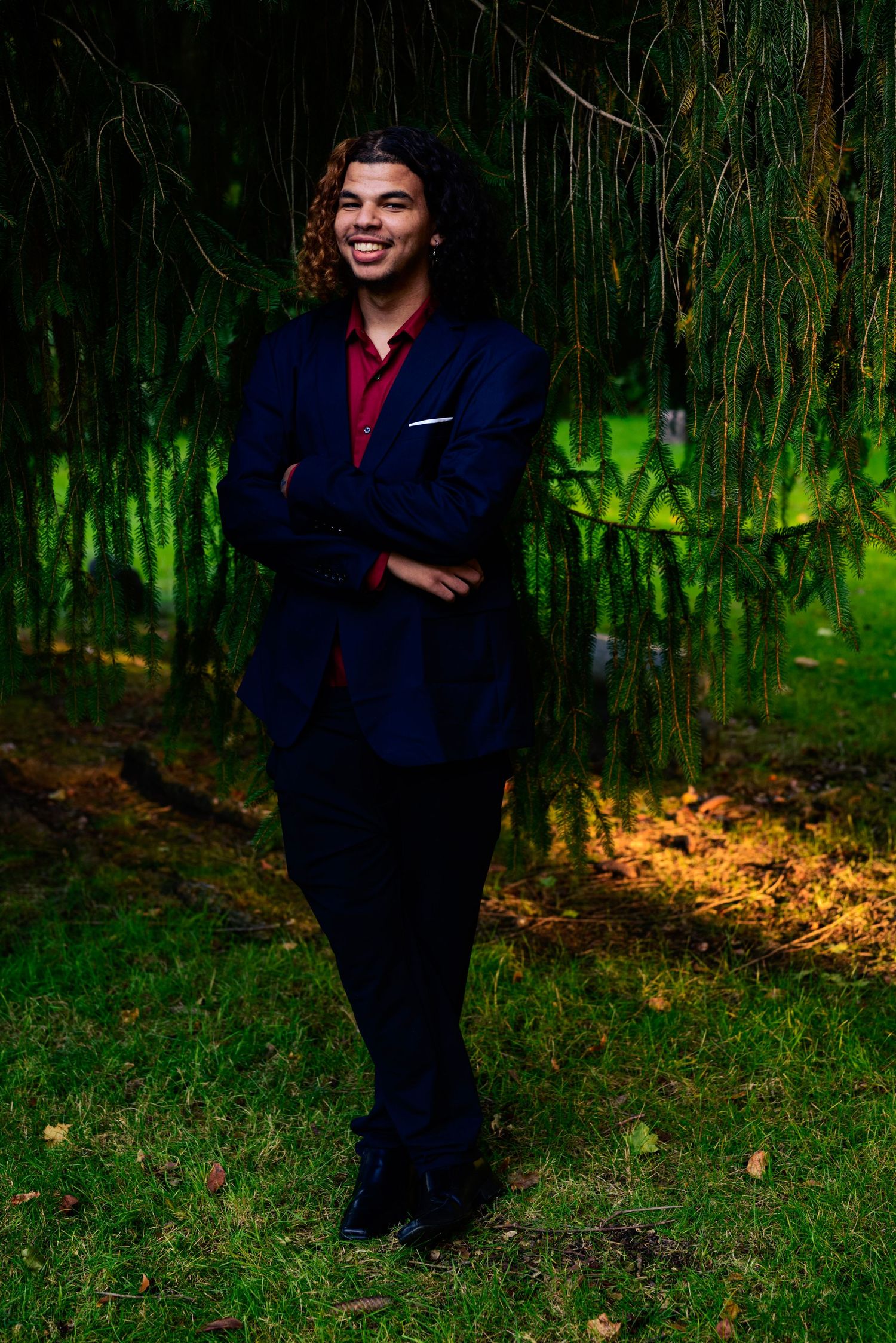 High school homecoming event. Young man in navy suit and red shirt smiling with arms crossed outdoors near weeping willow tree. NEO OHIO