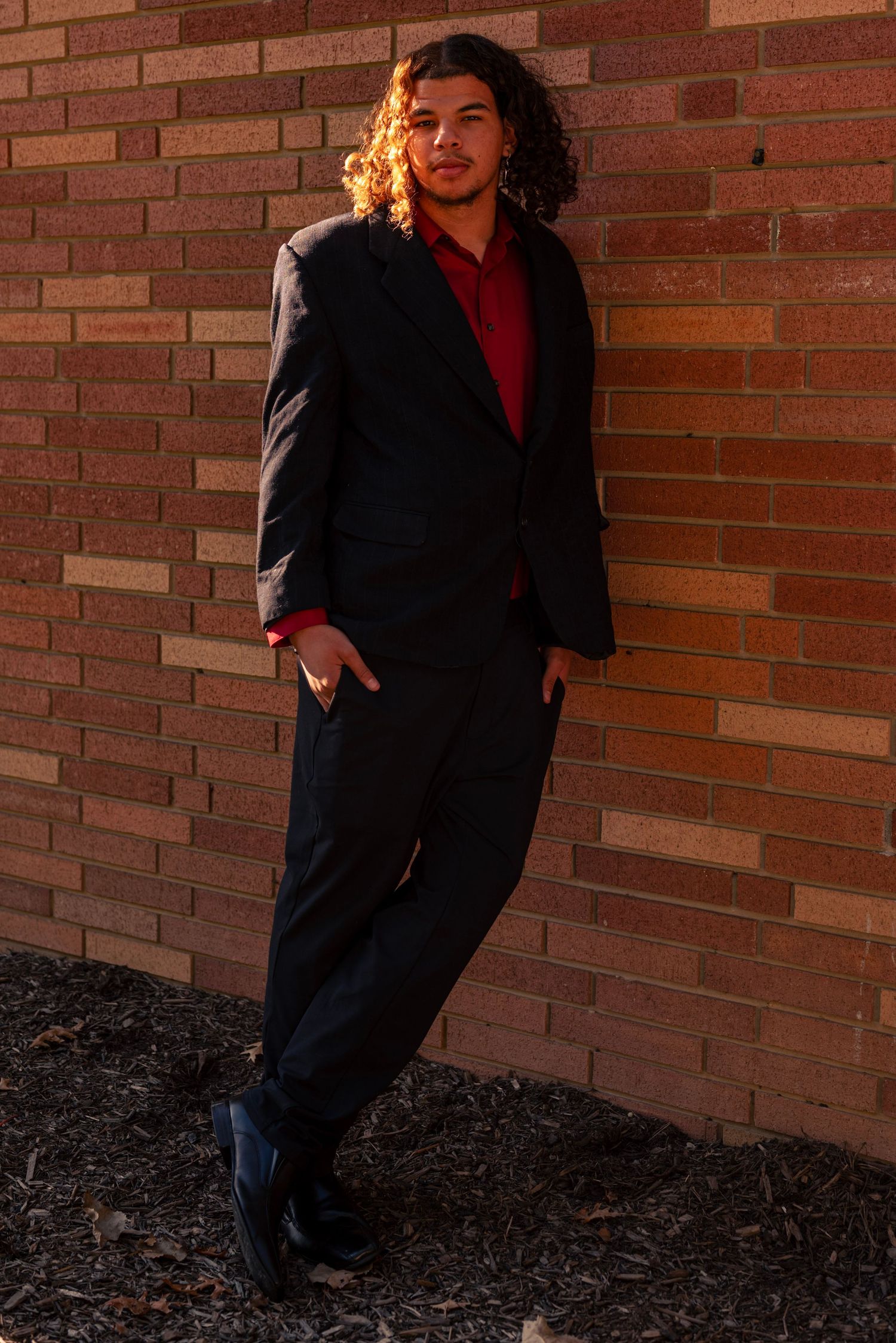 Young man with curly hair in black suit and red shirt leaning against brick wall in golden hour light. High School Homecoming event
