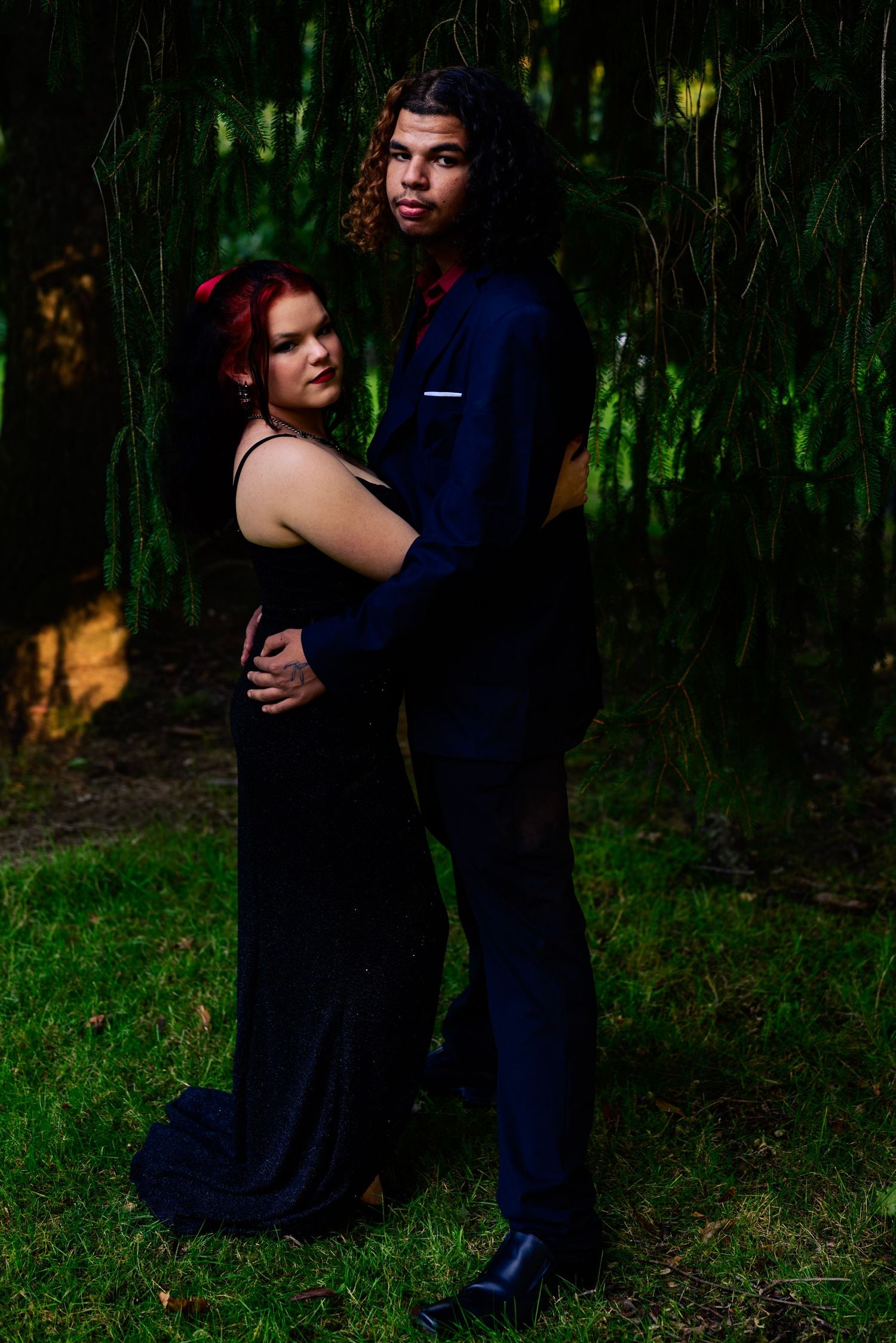A couple in formal attire, outdoors, a black gown, a navy suit, posed under weeping willow branches. High school homecoming event, NEO OH