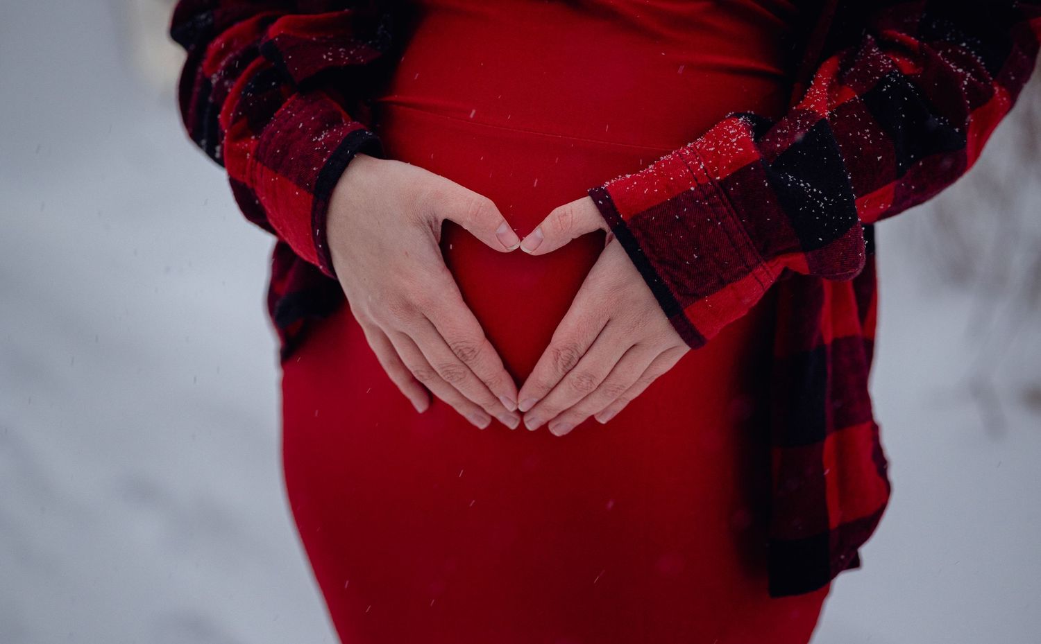 Pregnant woman in red dress forming heart shape with hands over baby bump in snowy winter setting. Neo Ohio, outdoors, maternity