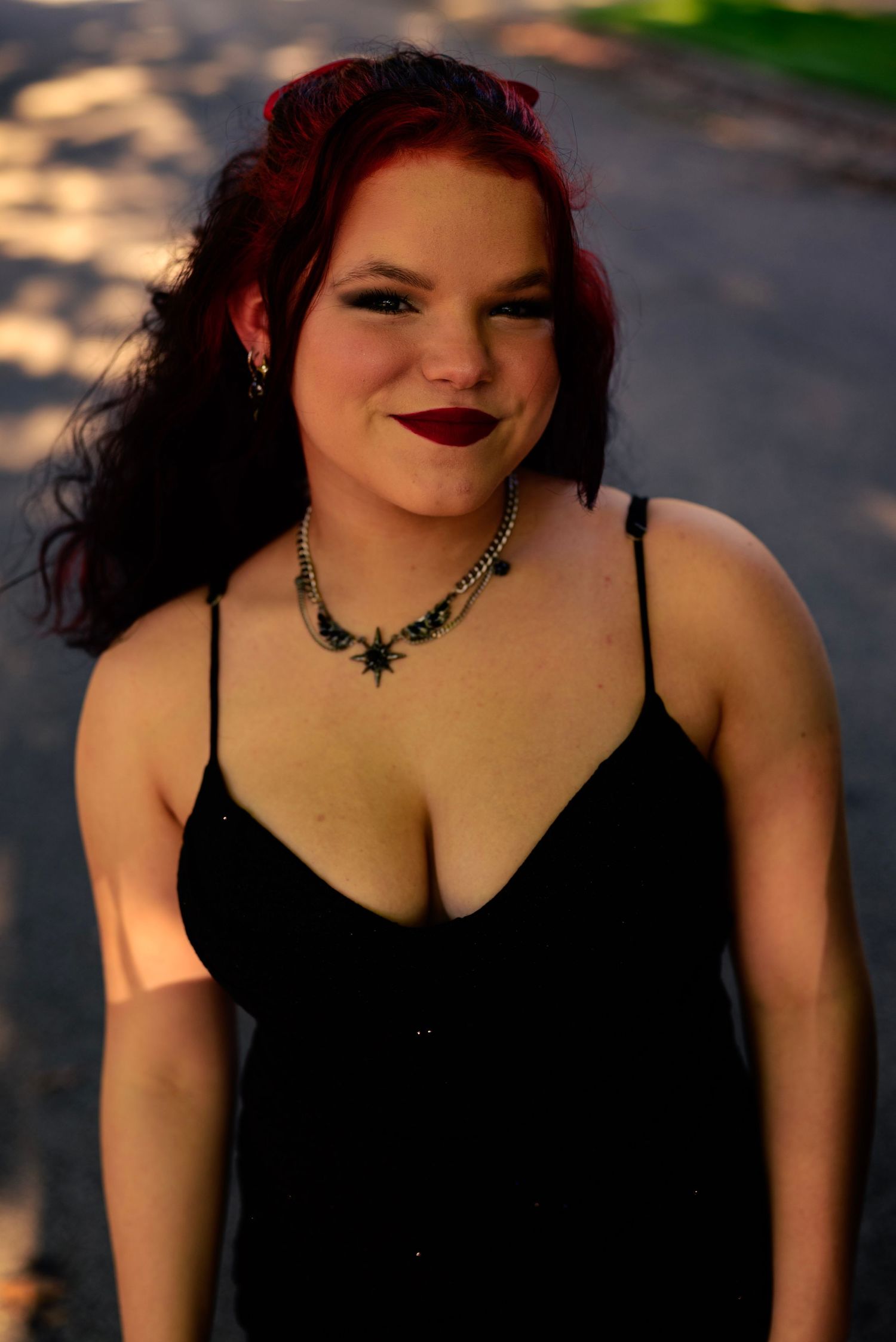 Young woman with red hair wearing a black dress and star necklace smiling outdoors in warm sunlight. High school Homecoming event