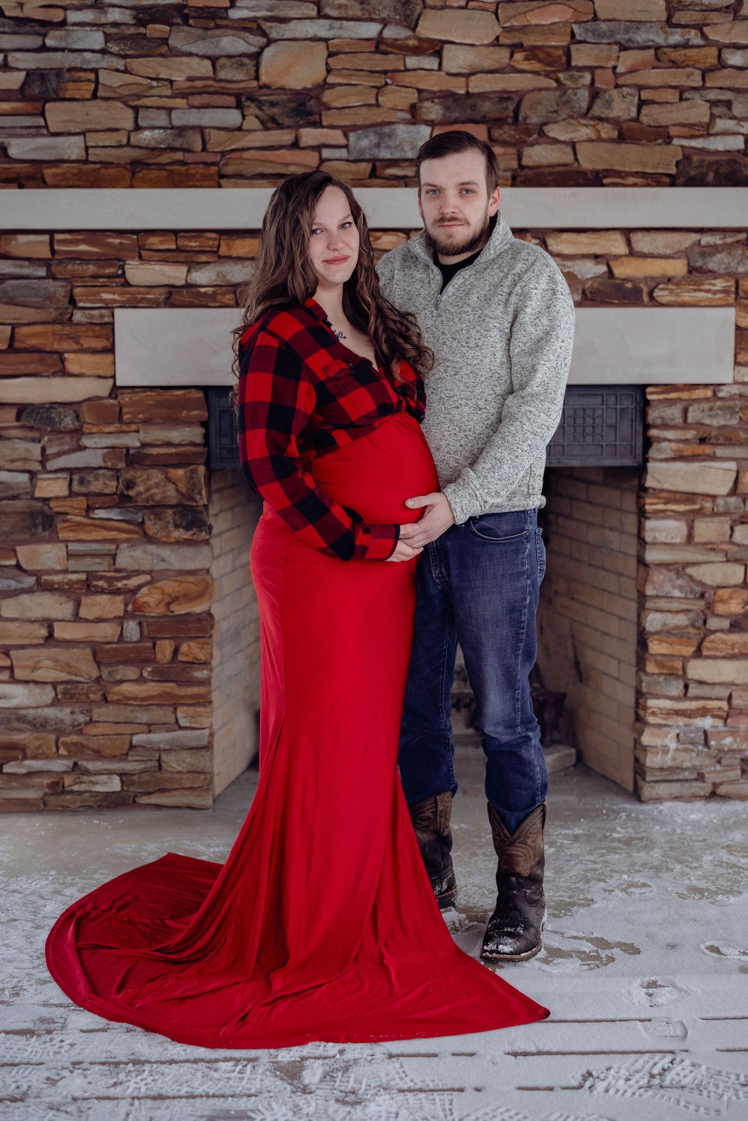 Pregnant couple poses in front of stone fireplace, woman in red  & plaid dress & man in gray sweater w/ jeans. Neo Ohio, outdoors, maternity