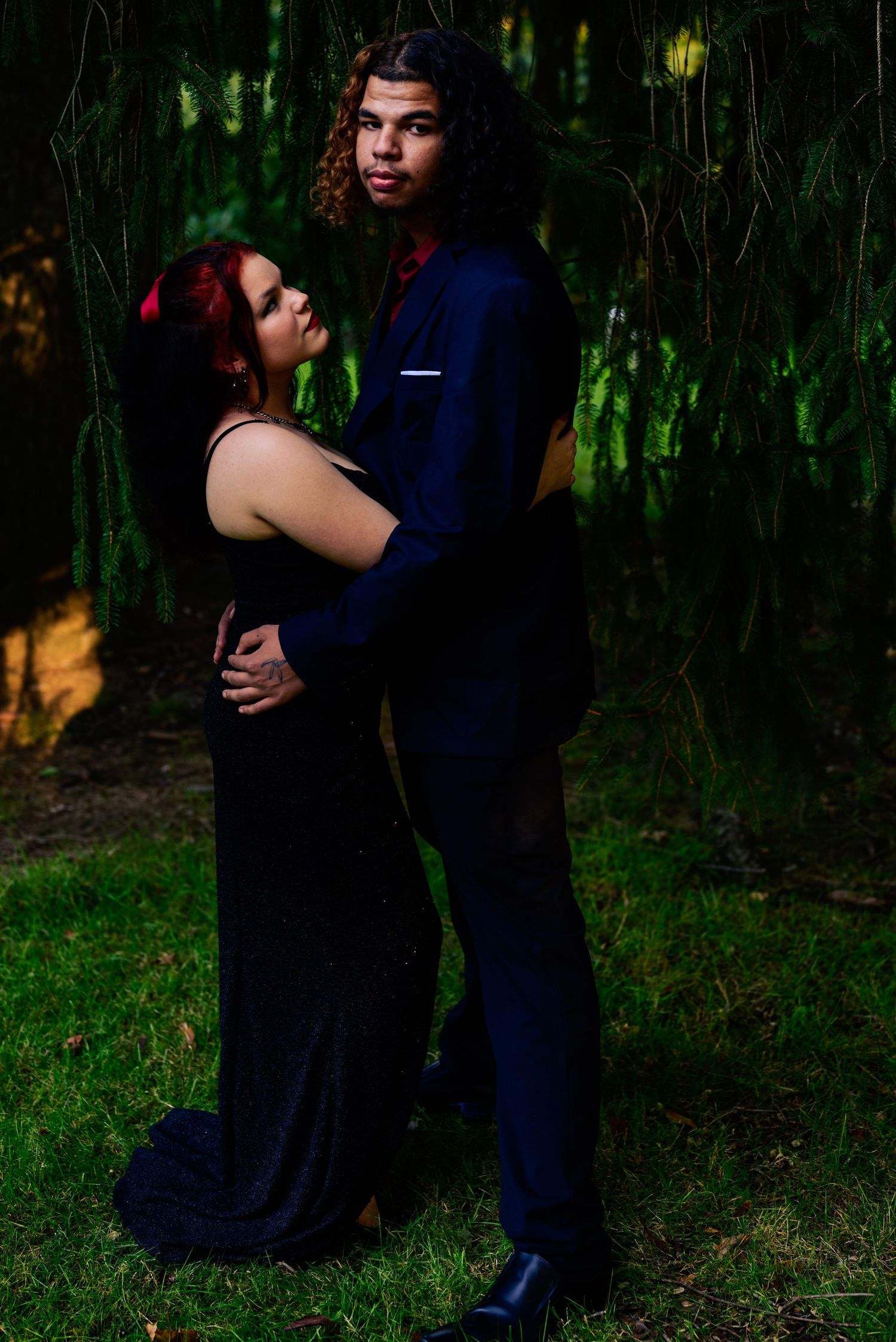 Ohio teen Couple in formal attire, outdoors, woman in black gown and man in navy suit among lush green trees. High school homecoming event.