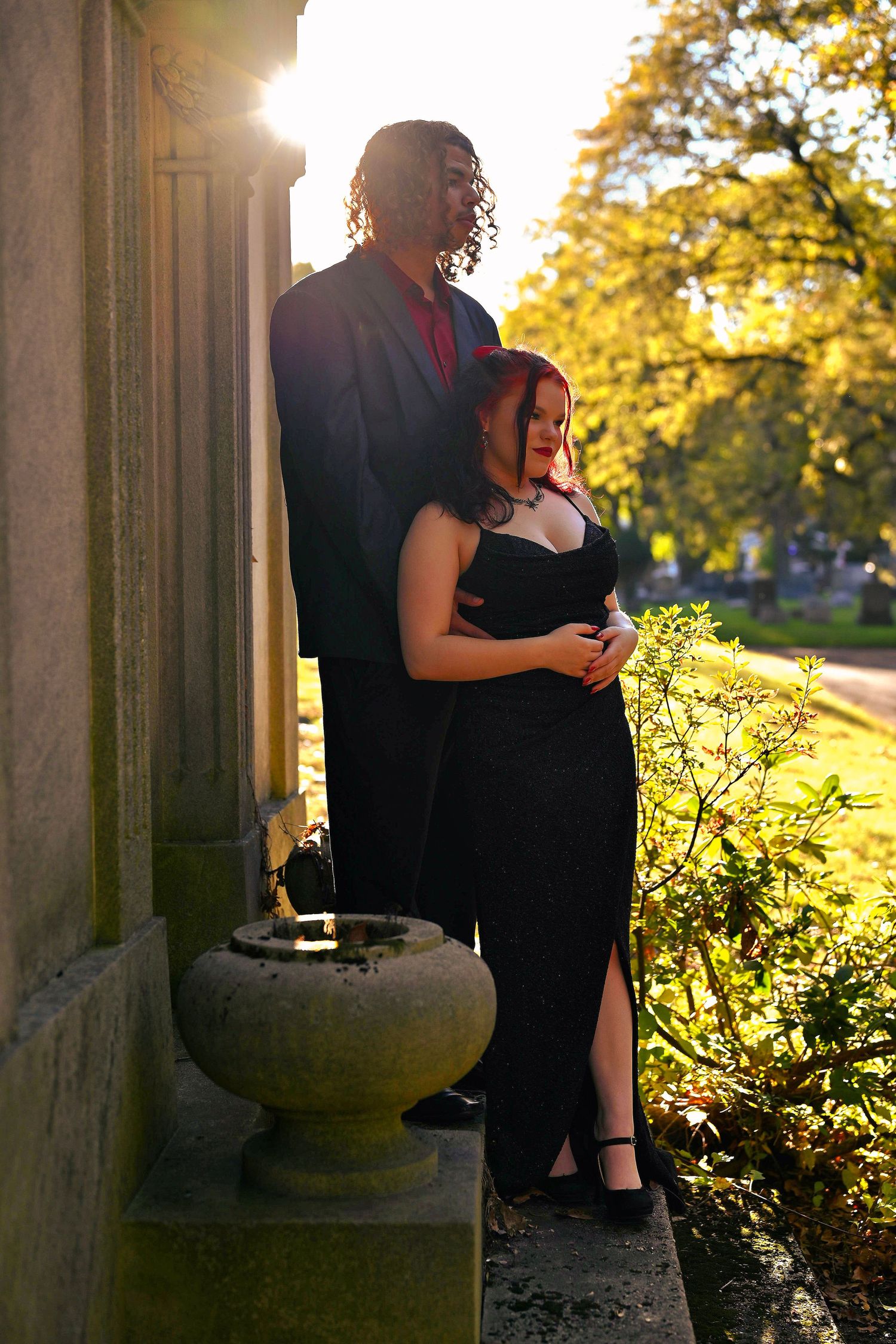 Couple in formal attire posing by stone column at sunset, woman in black gown, man in dark suit, lush green trees background.