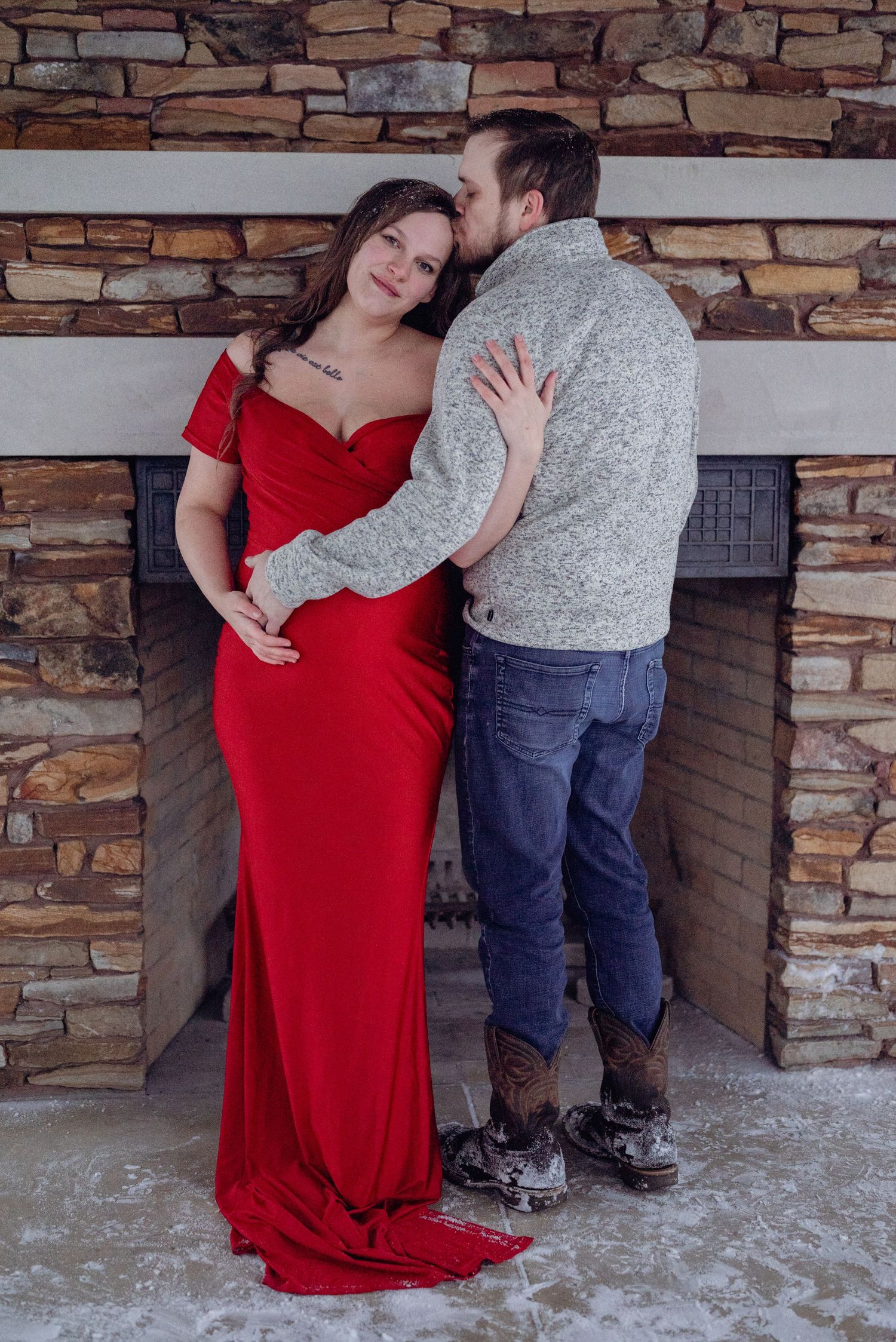 Pregnant woman in red gown poses with partner by stone fireplace in romantic maternity photo shoot. Neo Ohio, outdoors, maternity