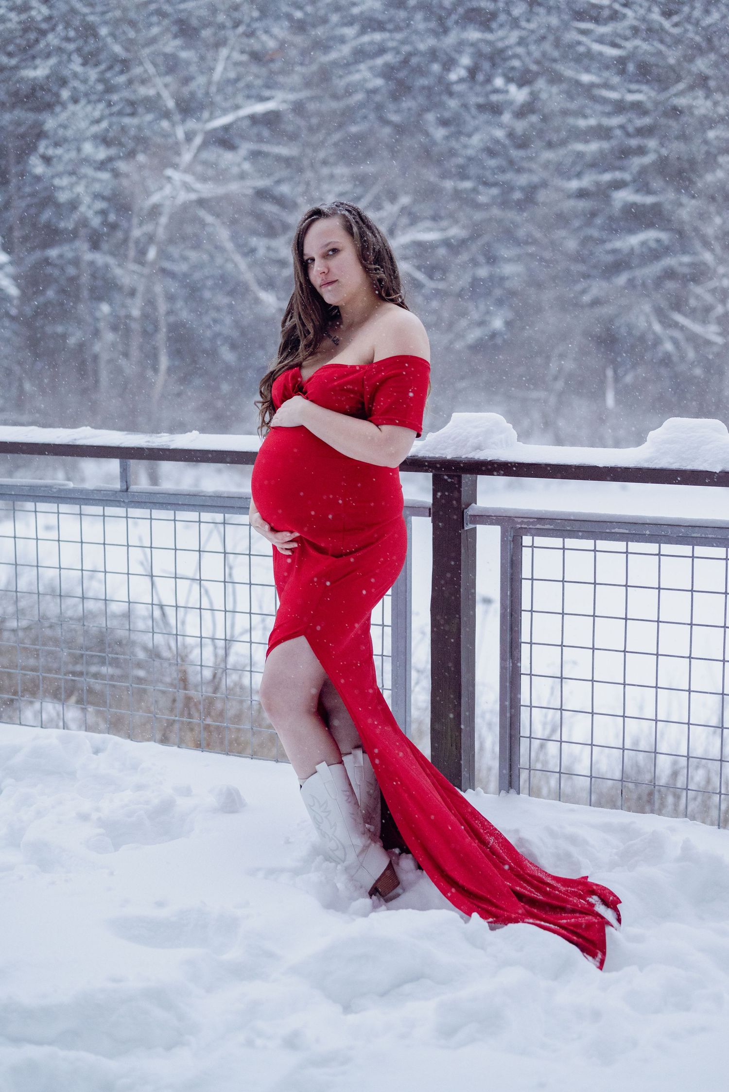 Pregnant woman in red off-shoulder gown & white boots poses in snowy winter landscape during maternity photo session. Ne Ohio, outdoors,