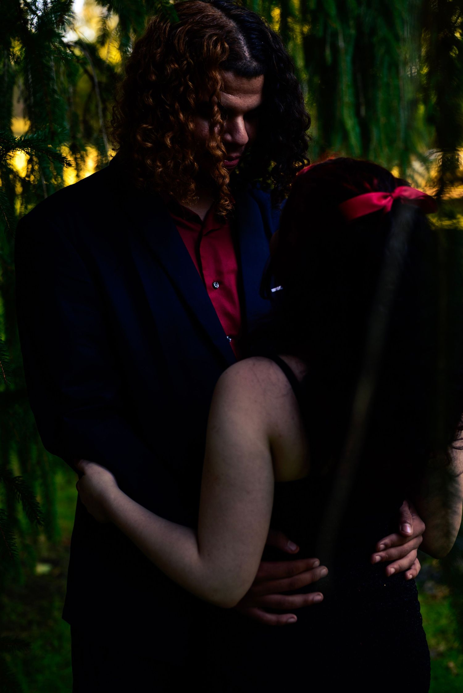 A couple embraces romantically in a dark forest setting, man in navy blazer & red shirt holding woman w/ red bow.  high school homecoming