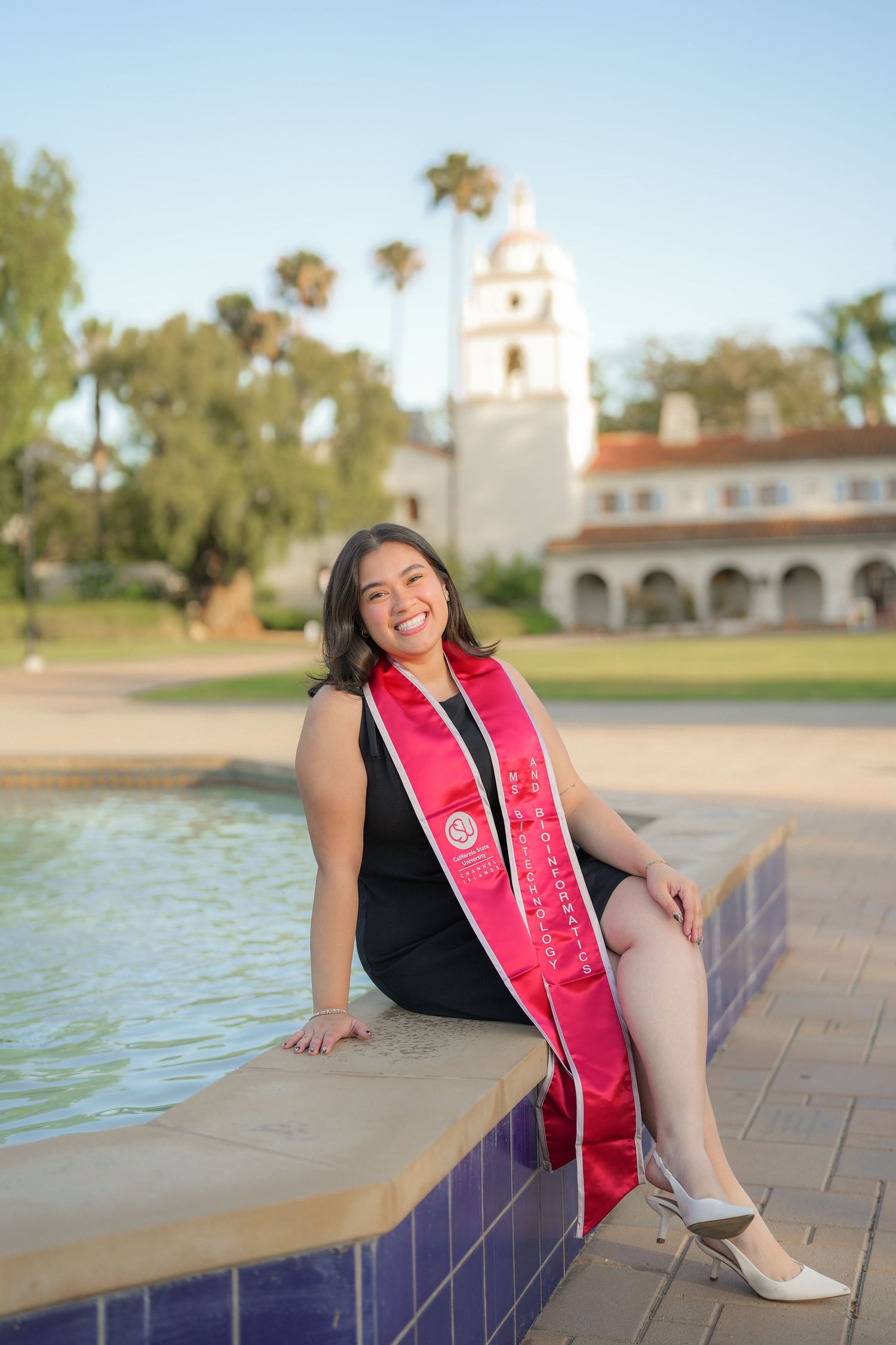 A smiling graduate in a black dress and red graduation sash sits on a tiled fountain ledge near a white bell tower.