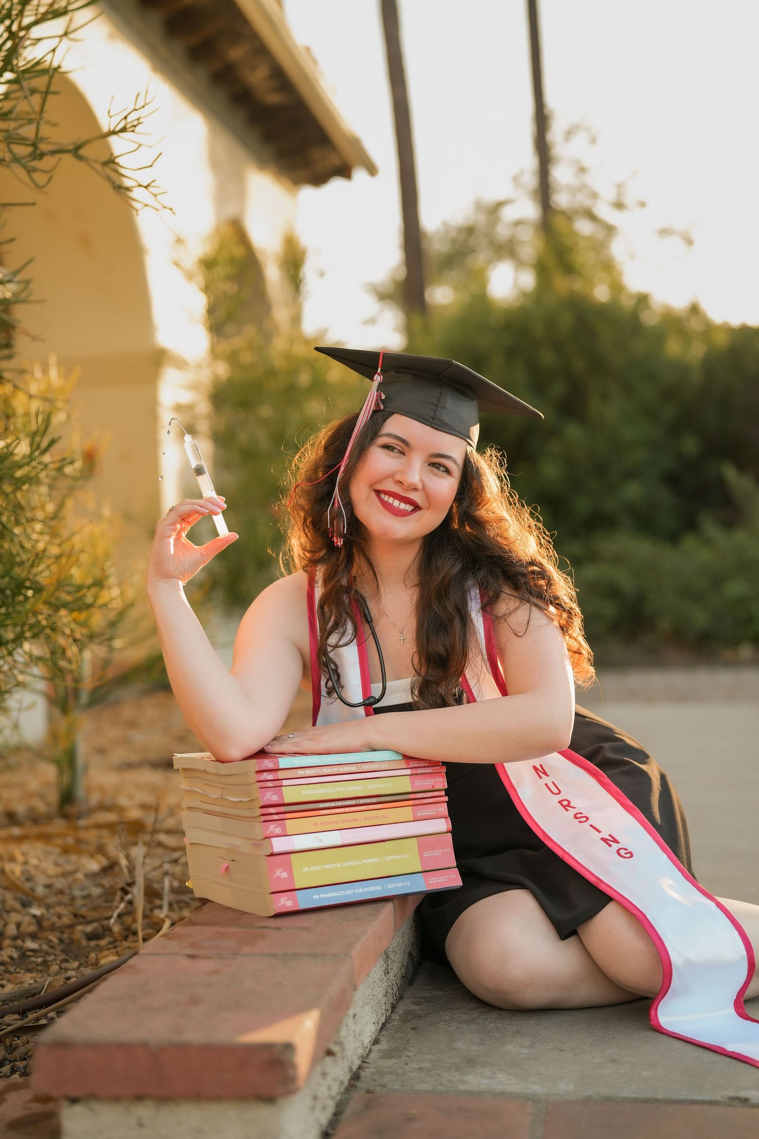 Smiling nursing graduate in cap and sash sits beside a stack of colorful books holding a small vial near a sunlit arched building.