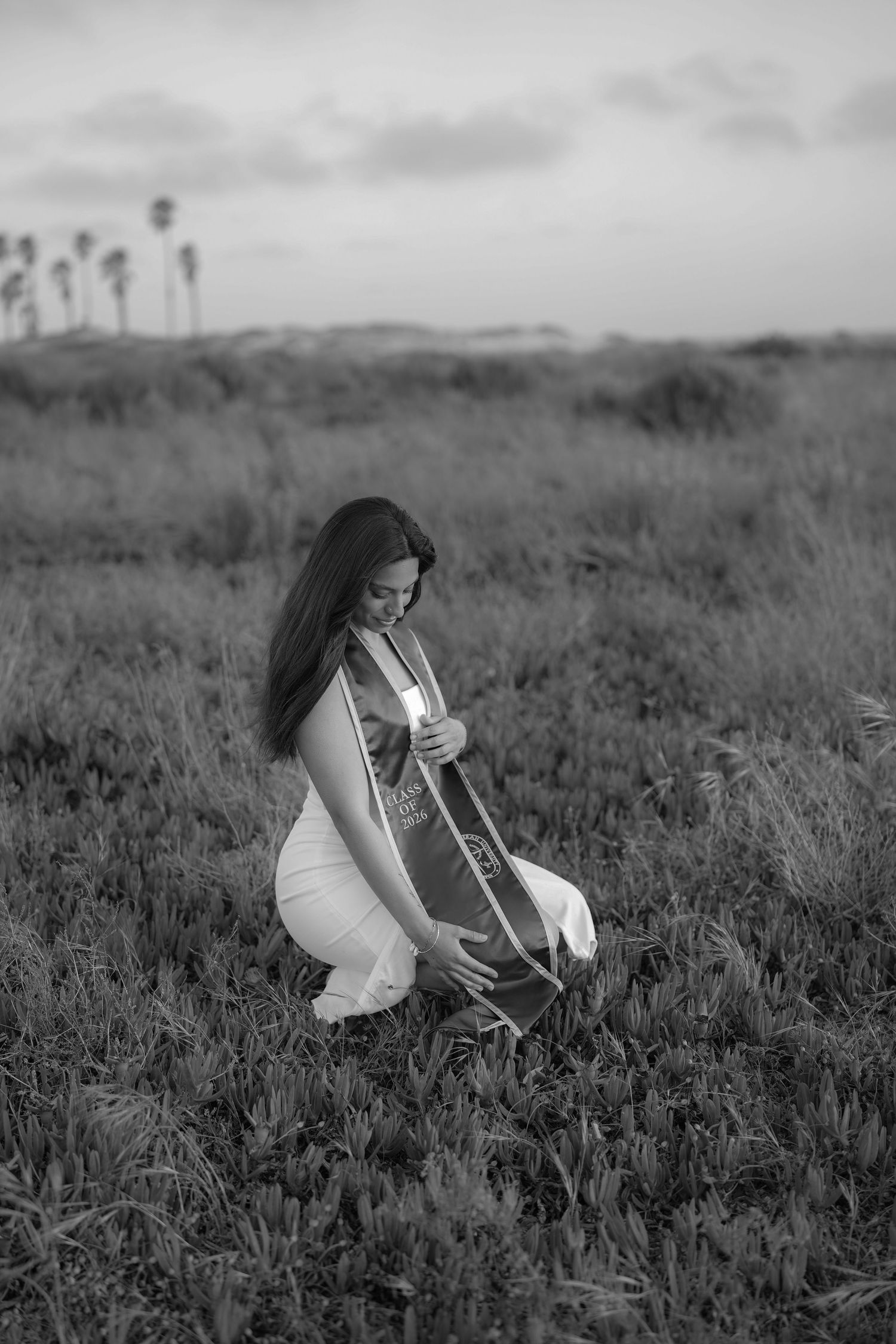 Black and white photo of a woman in a white dress kneeling in a grassy field while holding her graduation sash.