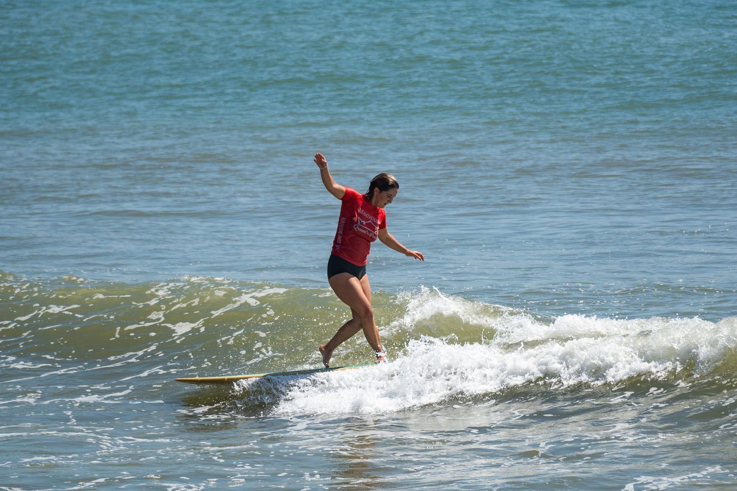 A surfer in a red shirt and black shorts balances with arms out on a longboard in foamy whitewater.