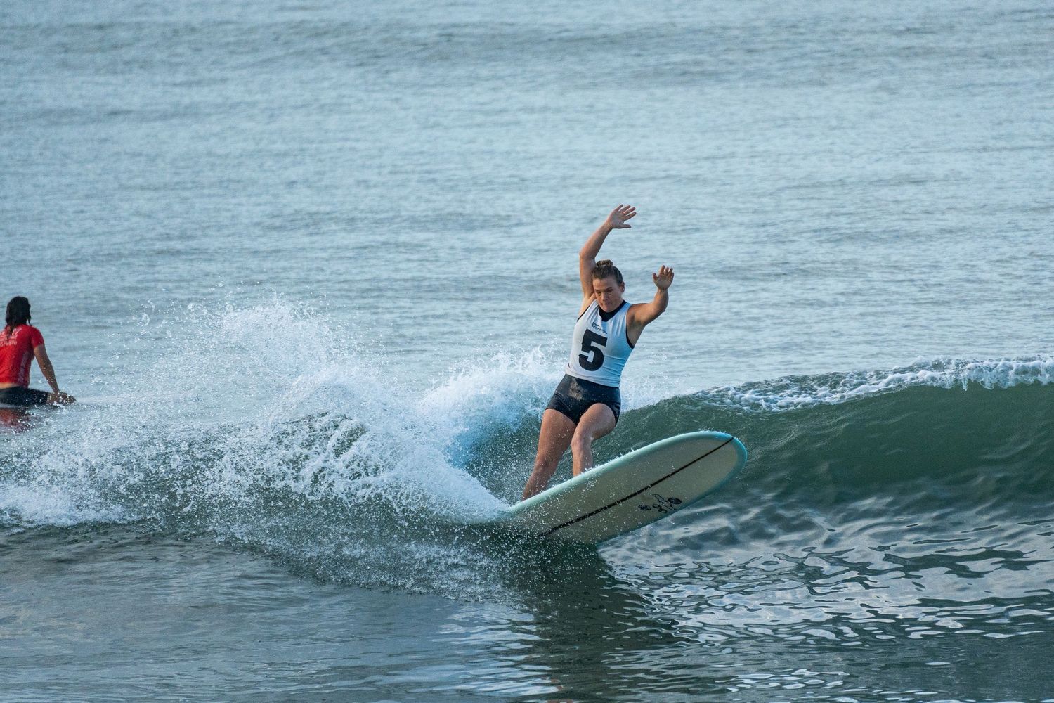 Competitive surfer in a numbered jersey raises her arms while riding a white longboard along a wave as another surfer paddles nearby.