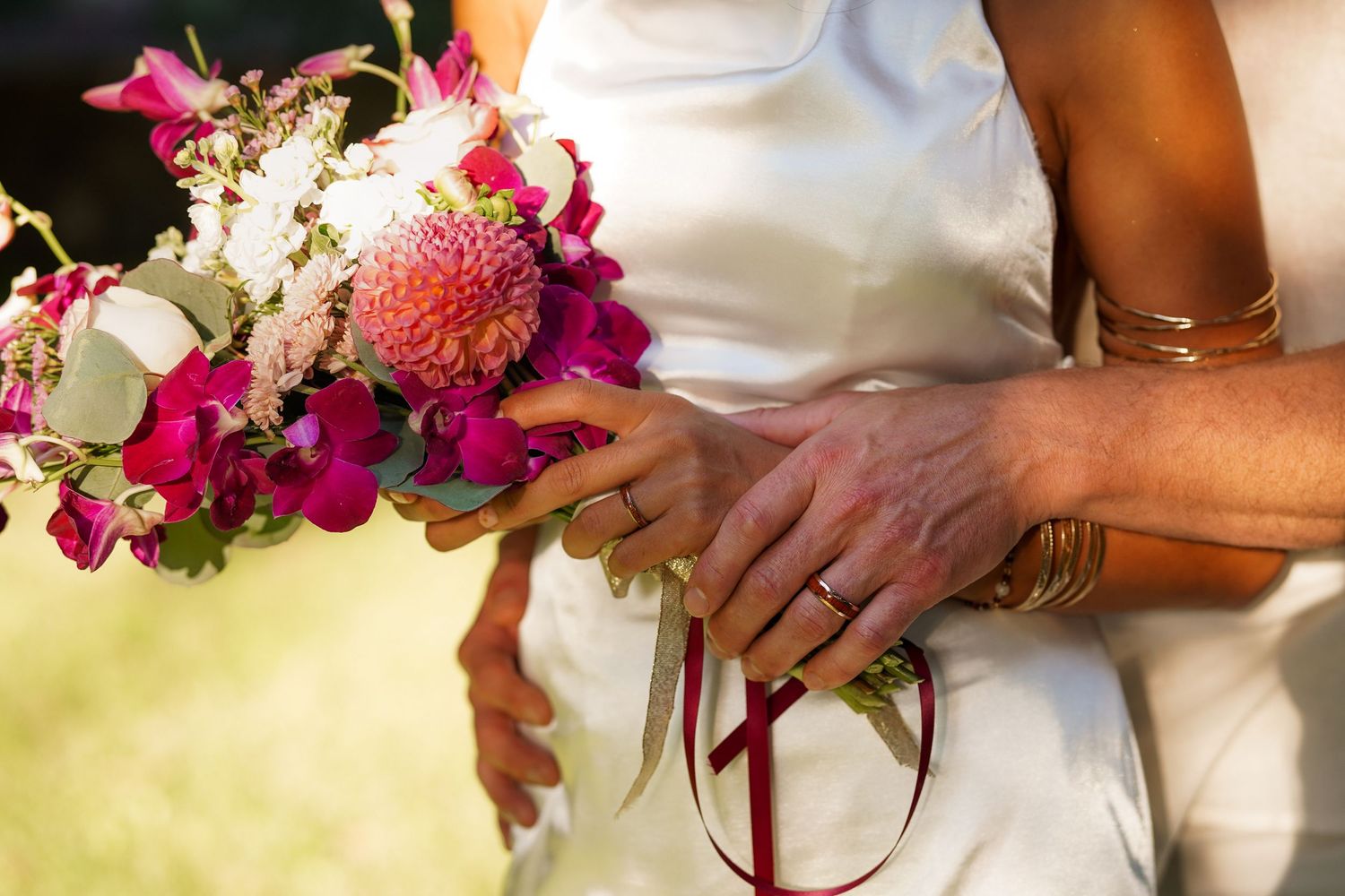Close-up of a bride and groom holding hands over a pink and white floral bouquet with a red ribbon, wearing gold bracelets and rings.
