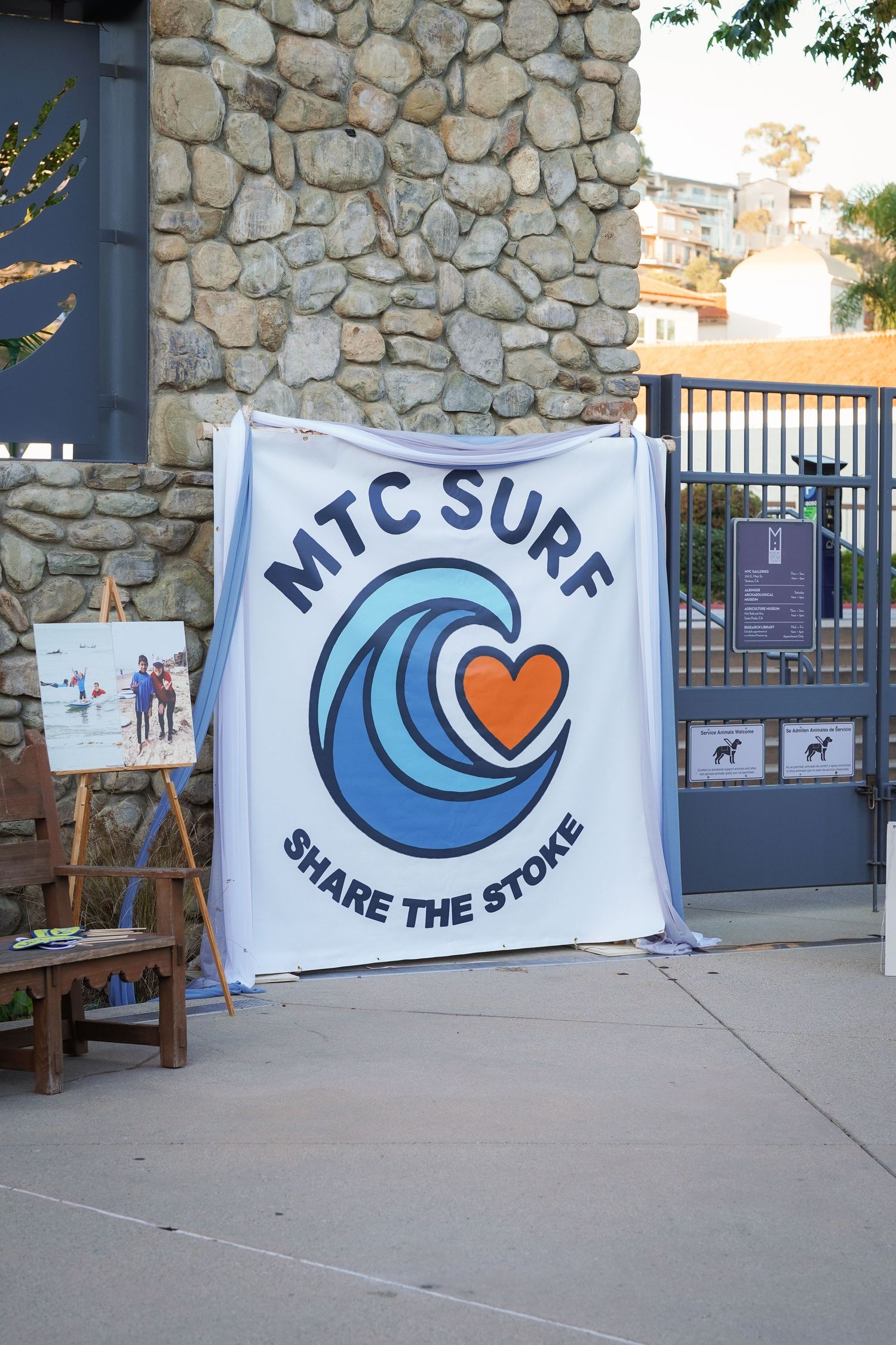 White banner reading MTC Surf Share the Stoke with a wave and orange heart logo hangs on a stone wall.