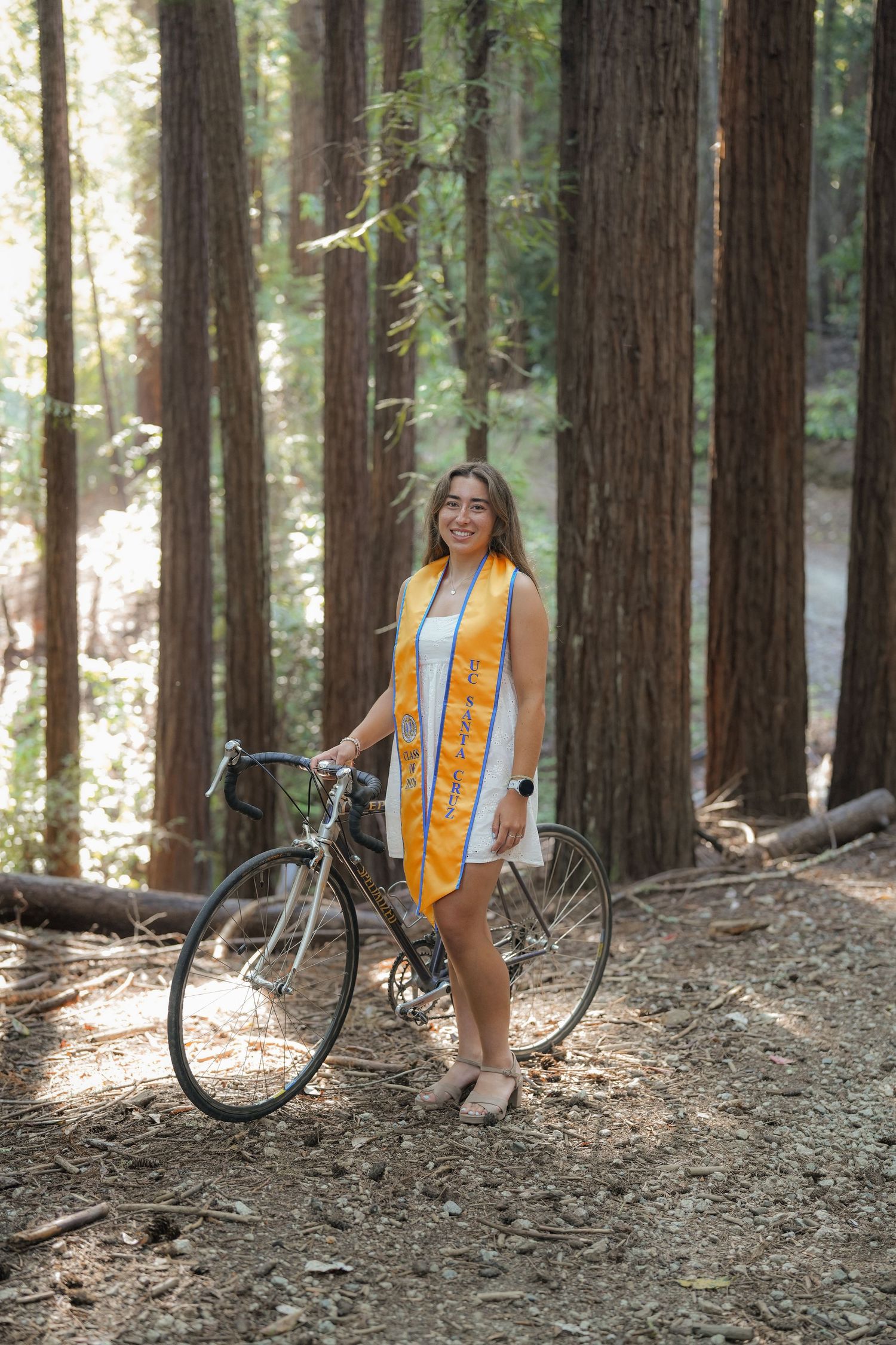 Graduate in a gold stole and white dress stands beside a road bike among tall redwood trees in a sunlit forest.